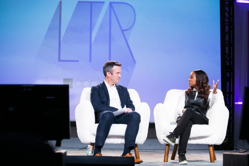 A man and a woman sit on white chairs during a discussion on stage, with a large logo in the background. The woman is gesturing as she speaks.