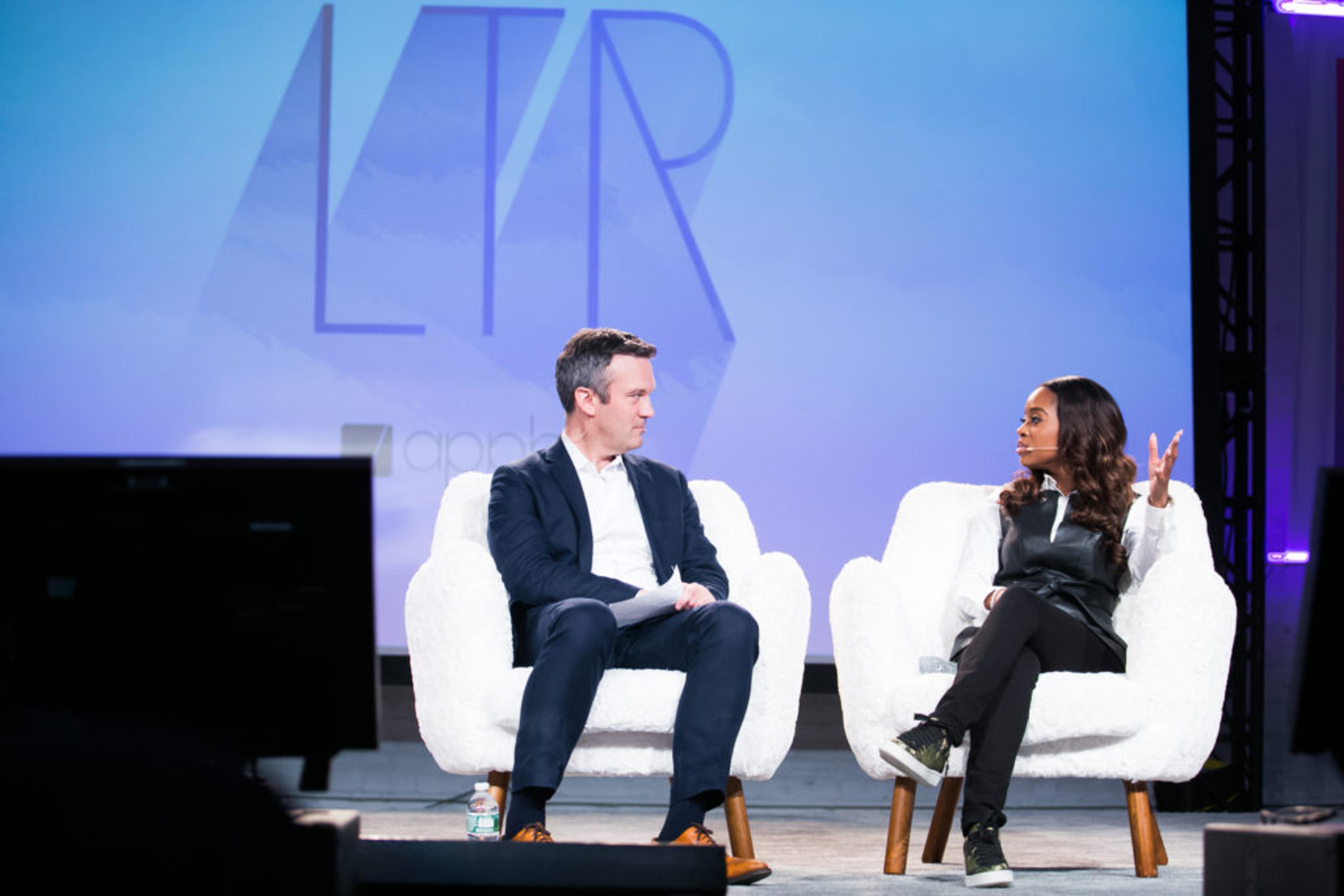 A man and a woman sit on white chairs during a discussion on stage, with a large logo in the background. The woman is gesturing as she speaks.
