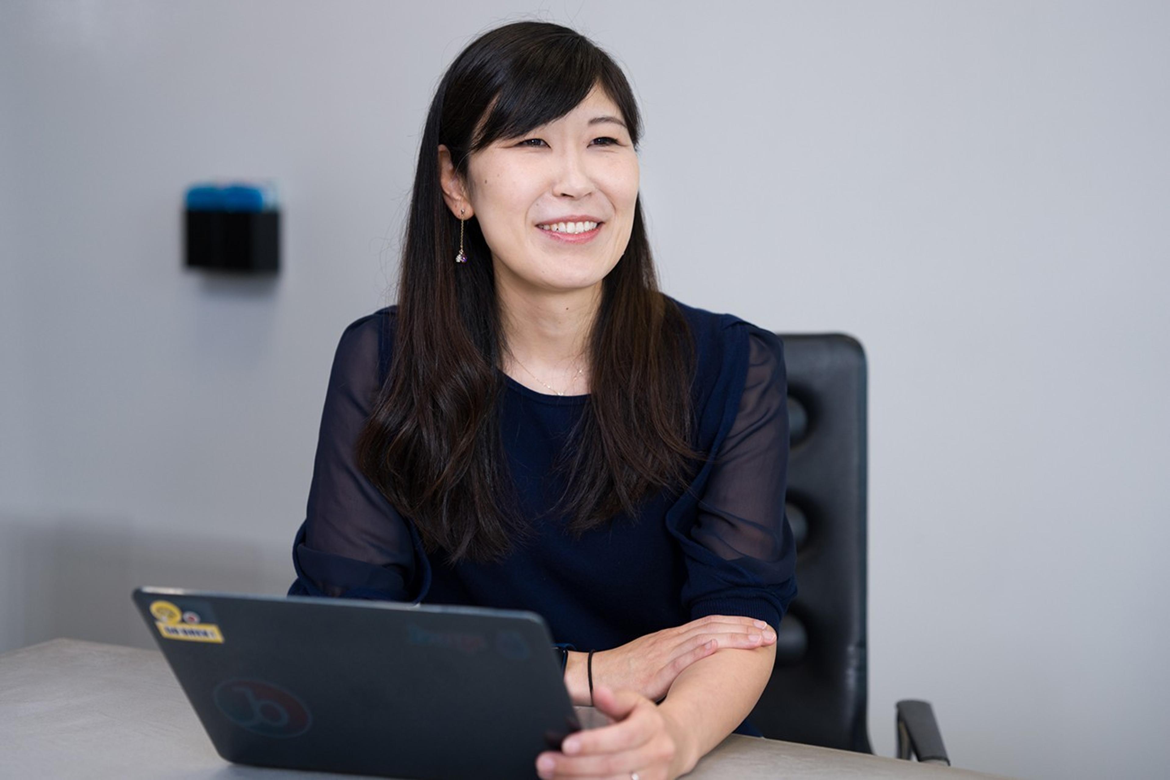 女性がノートパソコンの前で微笑んでいる。背景はシンプルなオフィス環境。