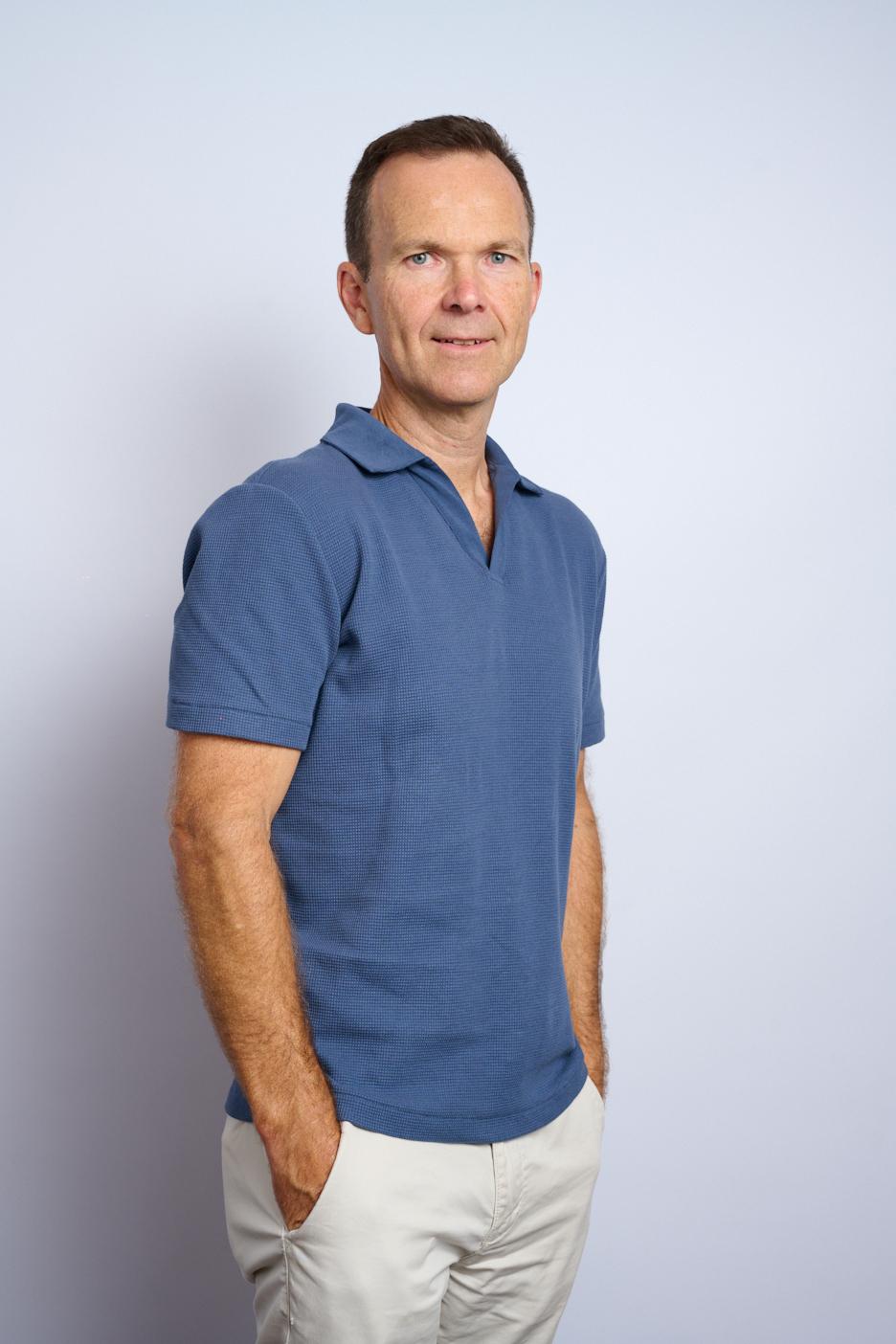Man standing against a plain light background, wearing a blue short-sleeve shirt and light-colored pants with his hands in his pockets.