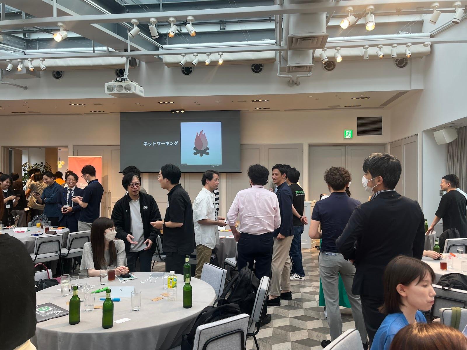 参加者がネットワーキングイベントで交流している様子。テーブルには飲み物が置かれ、スクリーンにはプレゼンテーションが映し出されている。