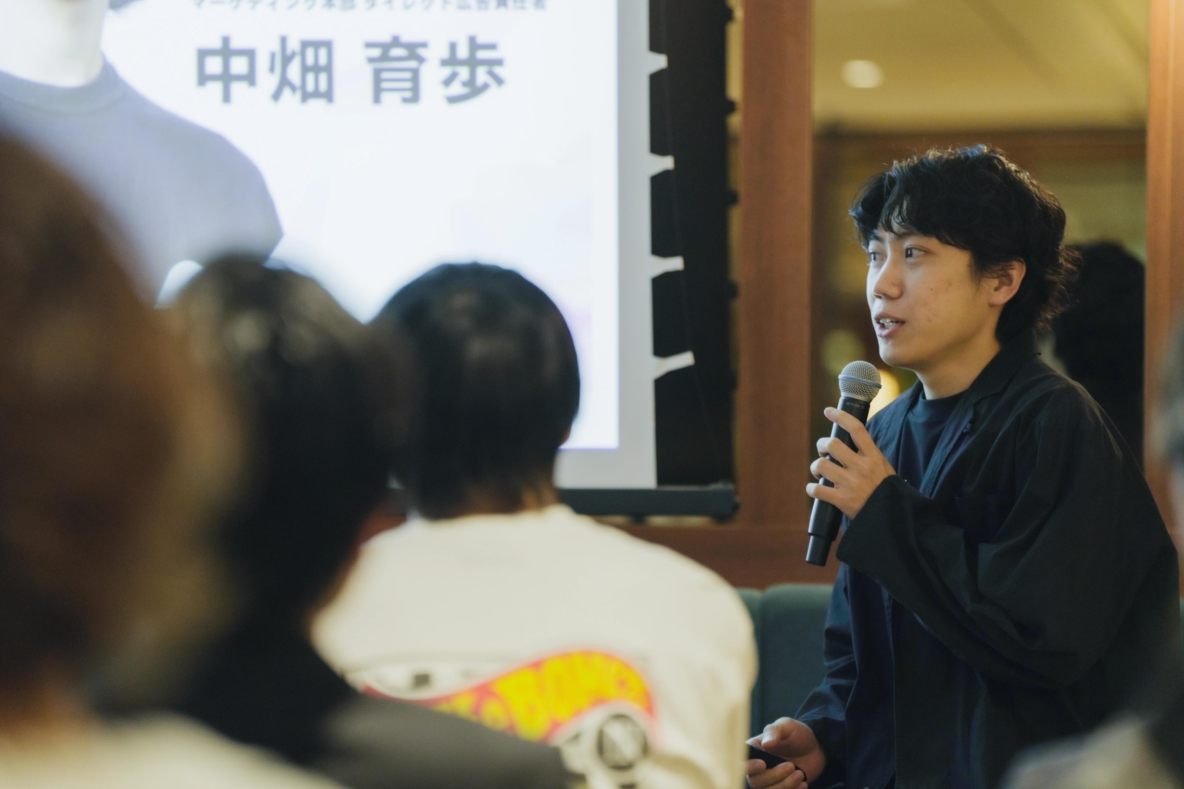 中畑育歩がマイクを持って話している様子。後ろには聴衆が座っている。