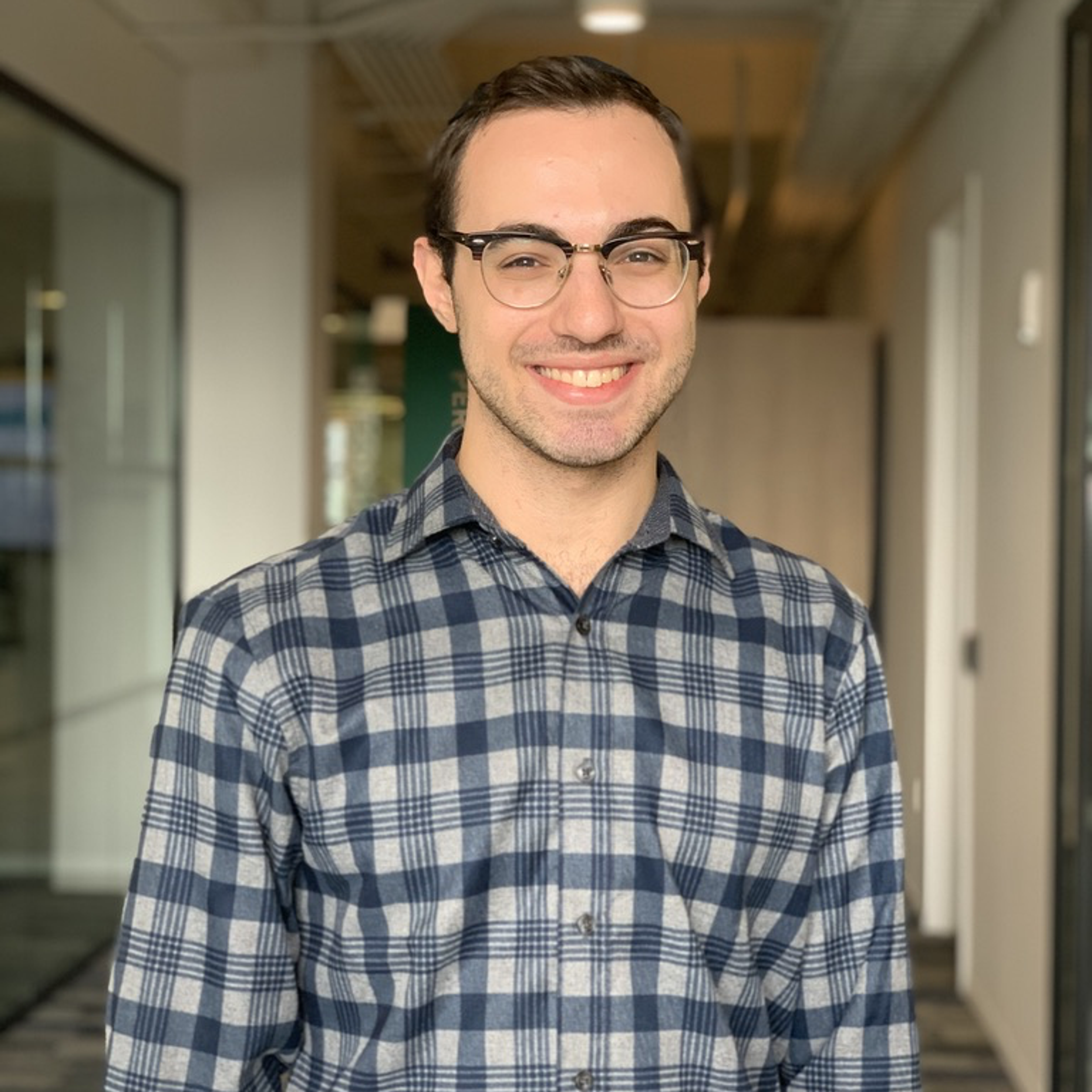 A young man with glasses smiles while standing in an office hallway. He is wearing a blue and gray plaid shirt.