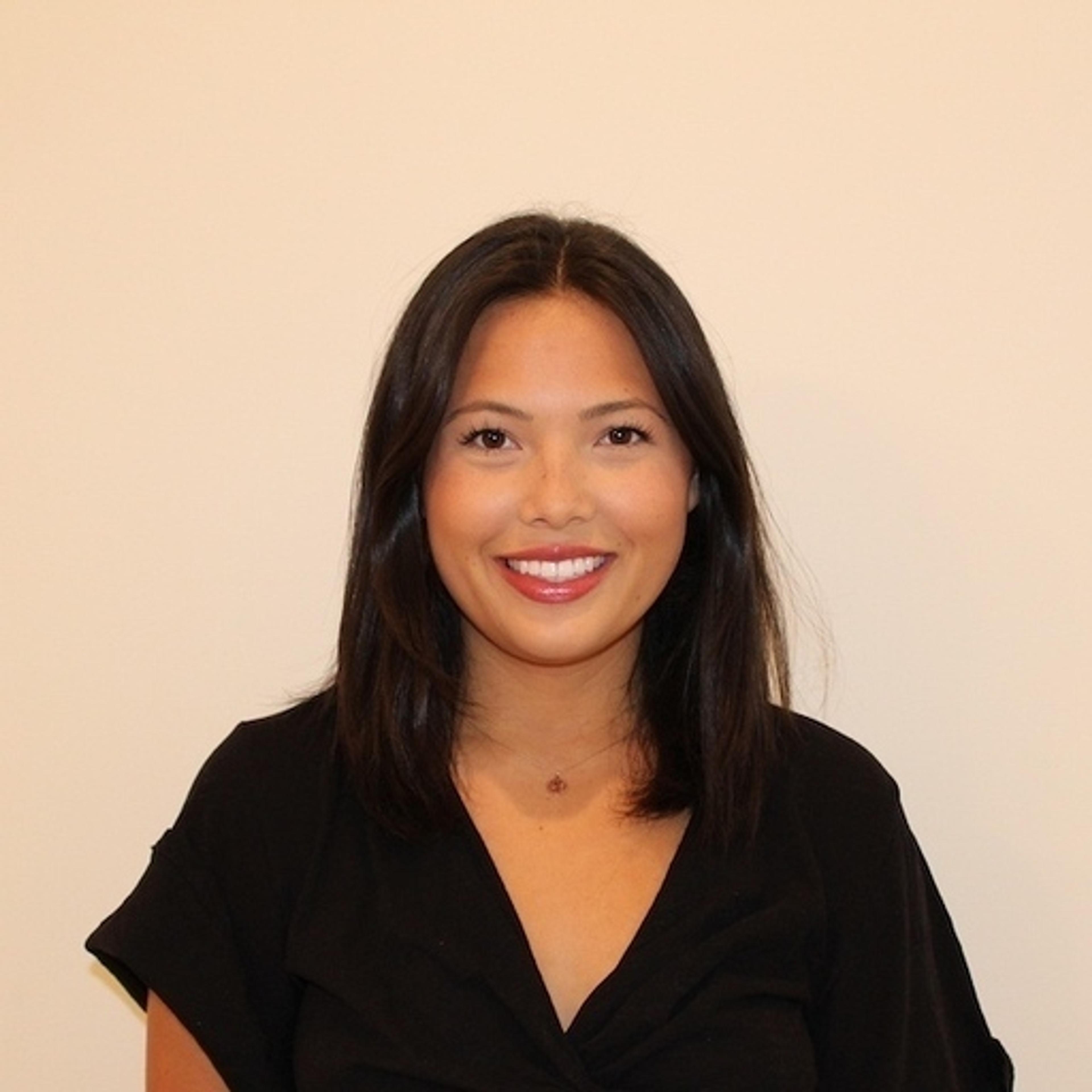 A woman with long, dark hair smiles at the camera, wearing a black top against a plain background.