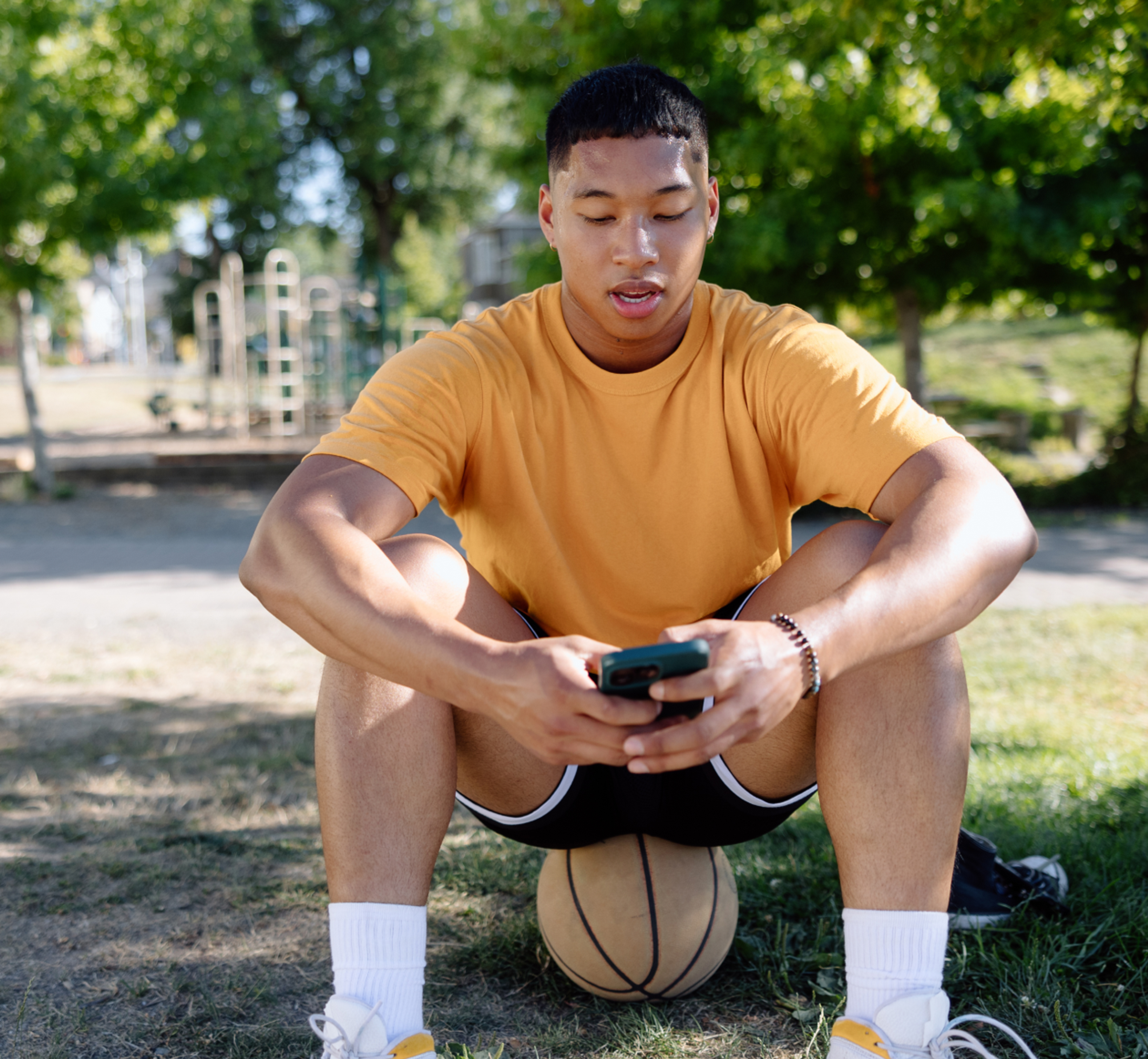 Young man in a yellow shirt sits outdoors on a basketball, looking at his phone in a park.