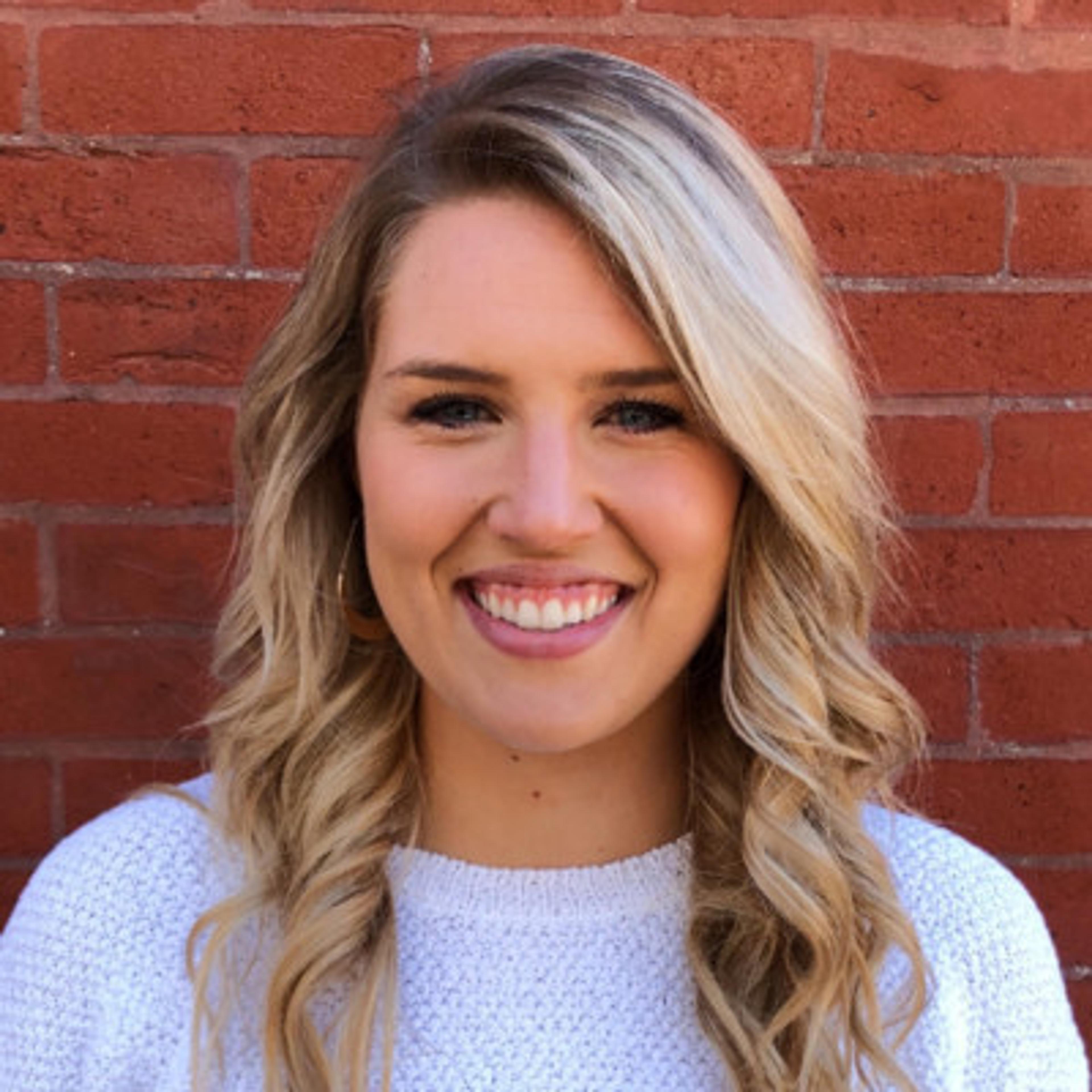 A smiling woman with long, wavy blonde hair stands in front of a brick wall. She is wearing a white sweater and has hoop earrings.