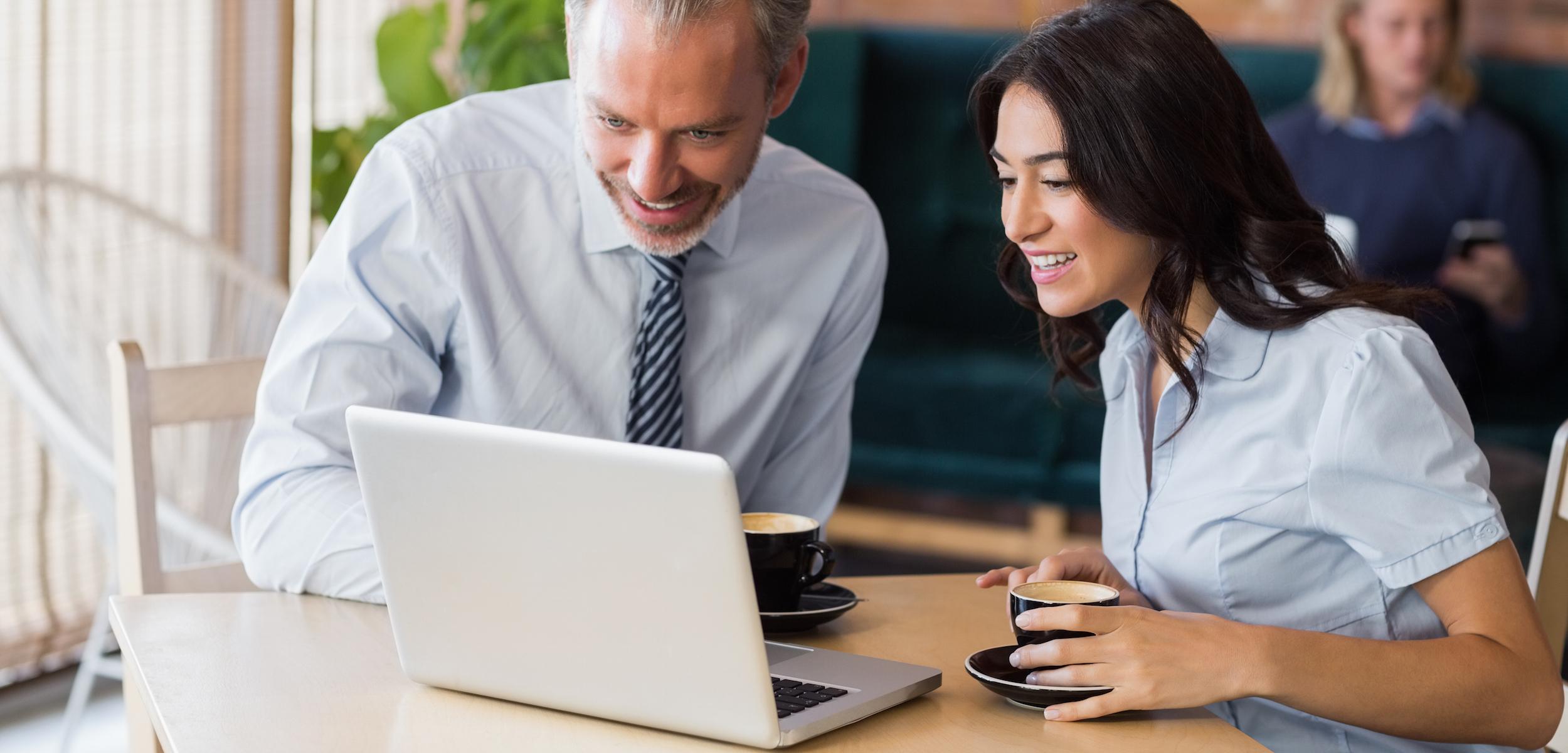 A man and a woman are sitting at a table, smiling and looking at a laptop while holding coffee cups. A person is visible in the background, engaged with their phone.