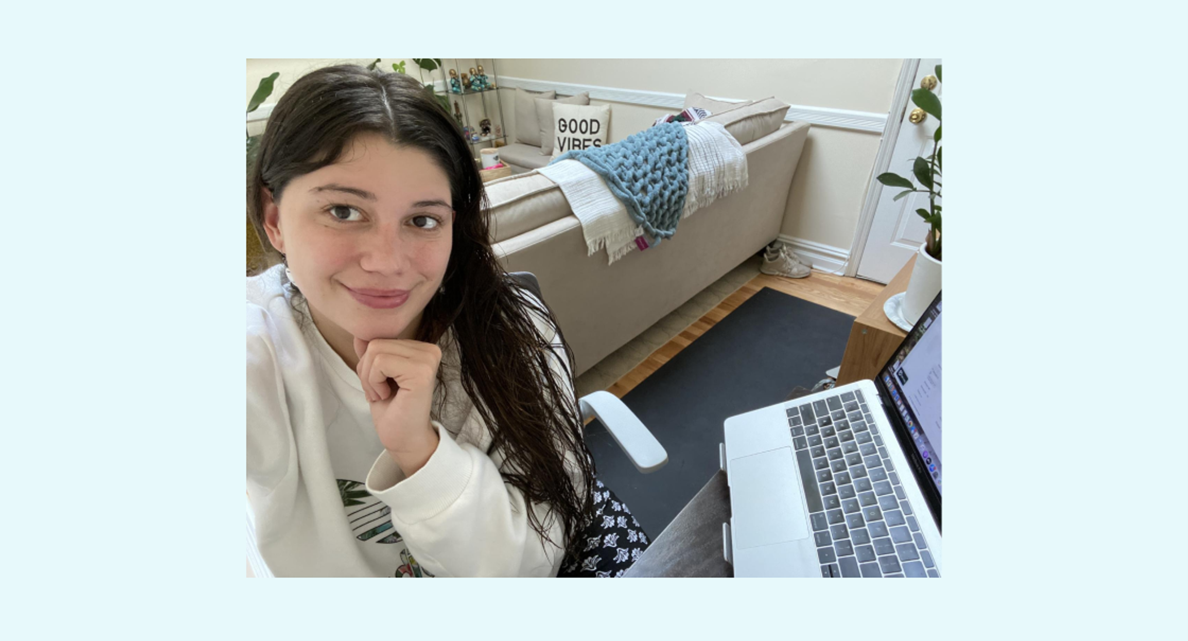 A young woman with long dark hair smiles at the camera while sitting at a desk with a laptop. In the background, there is a couch and decorative plants.