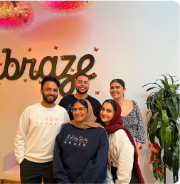 Five smiling people pose together indoors beneath a “brio” wall sign, with a tall leafy plant beside them.