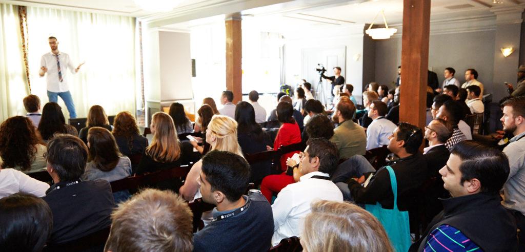 A speaker presents to an audience in a conference setting, with attendees seated and focused on the stage. Cameras capture the event from various angles.