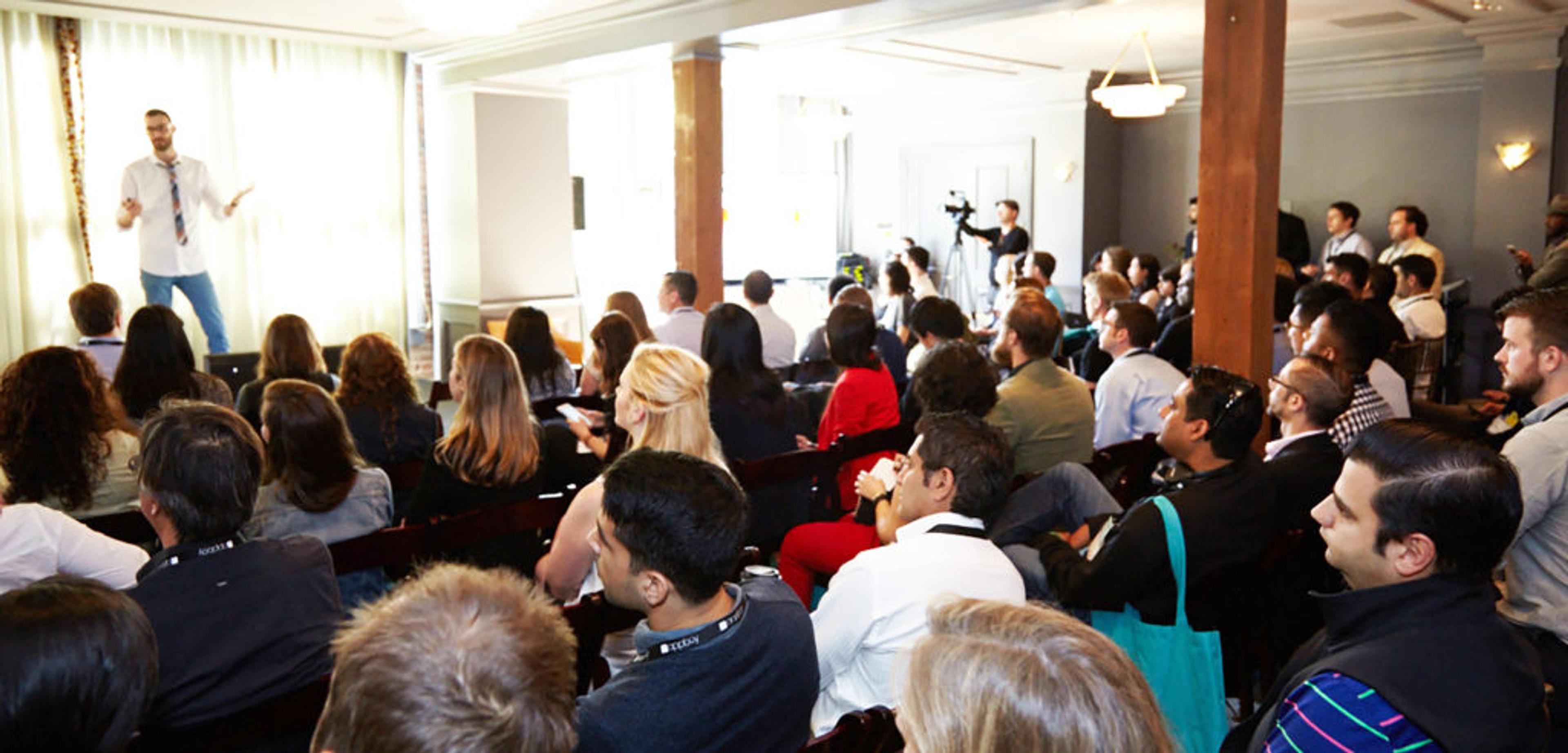 A speaker presents to an audience in a conference setting, with attendees seated and focused on the stage. Cameras capture the event from various angles.