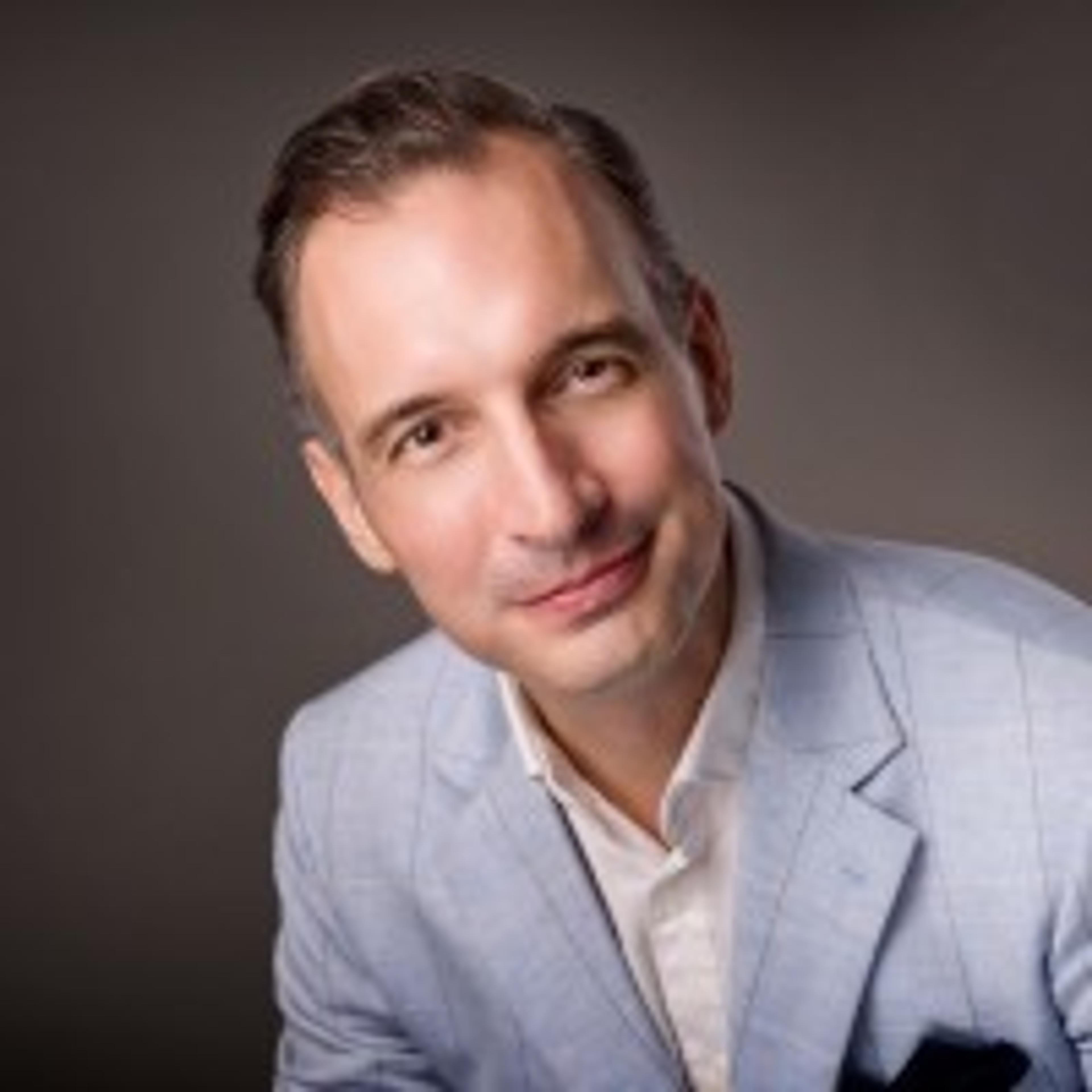 A man with short hair and a slight smile is wearing a light blue blazer over a white shirt. The background is a soft gray.