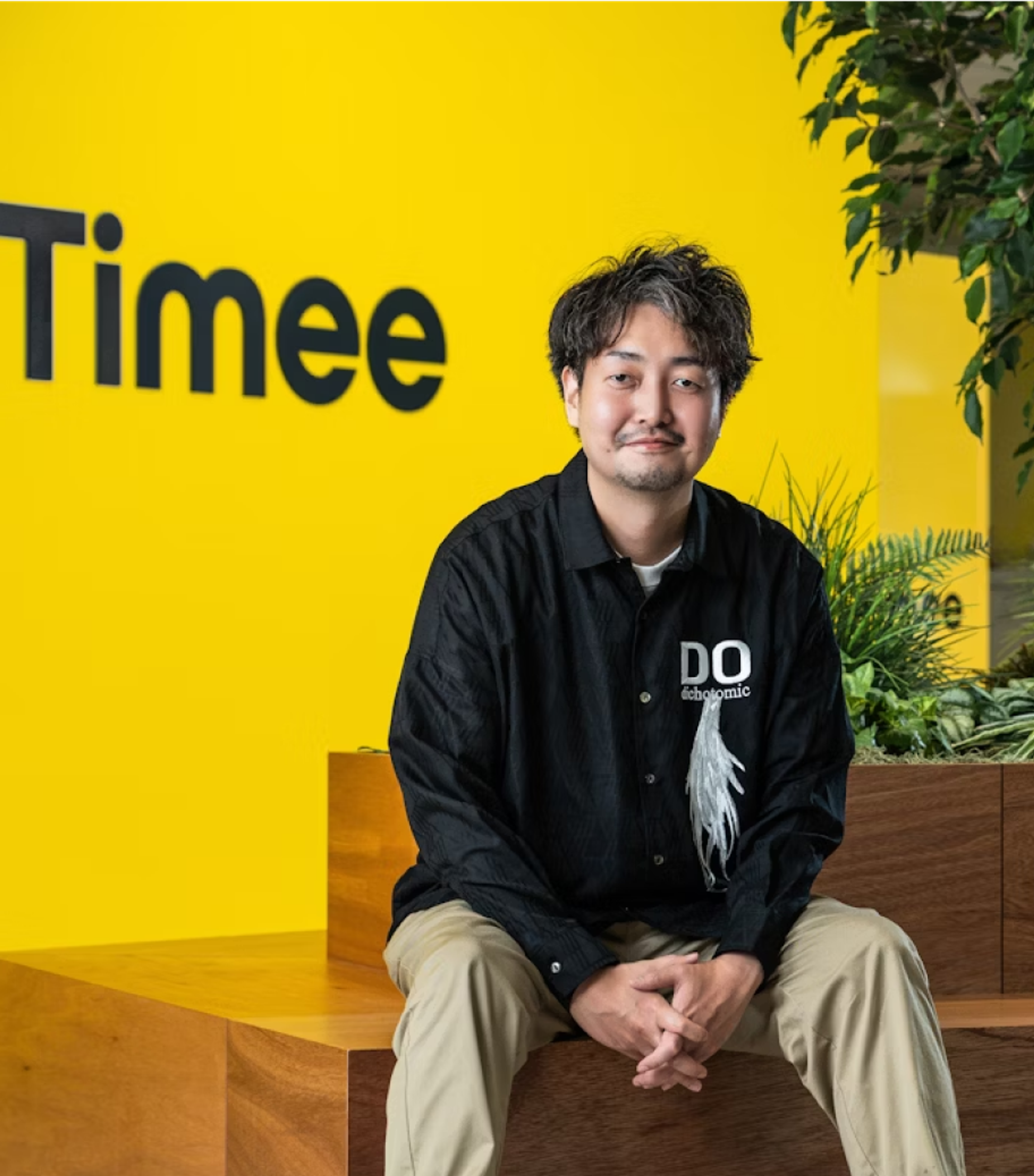 Timeeのロゴと黄色い壁の前で男性が座っている
