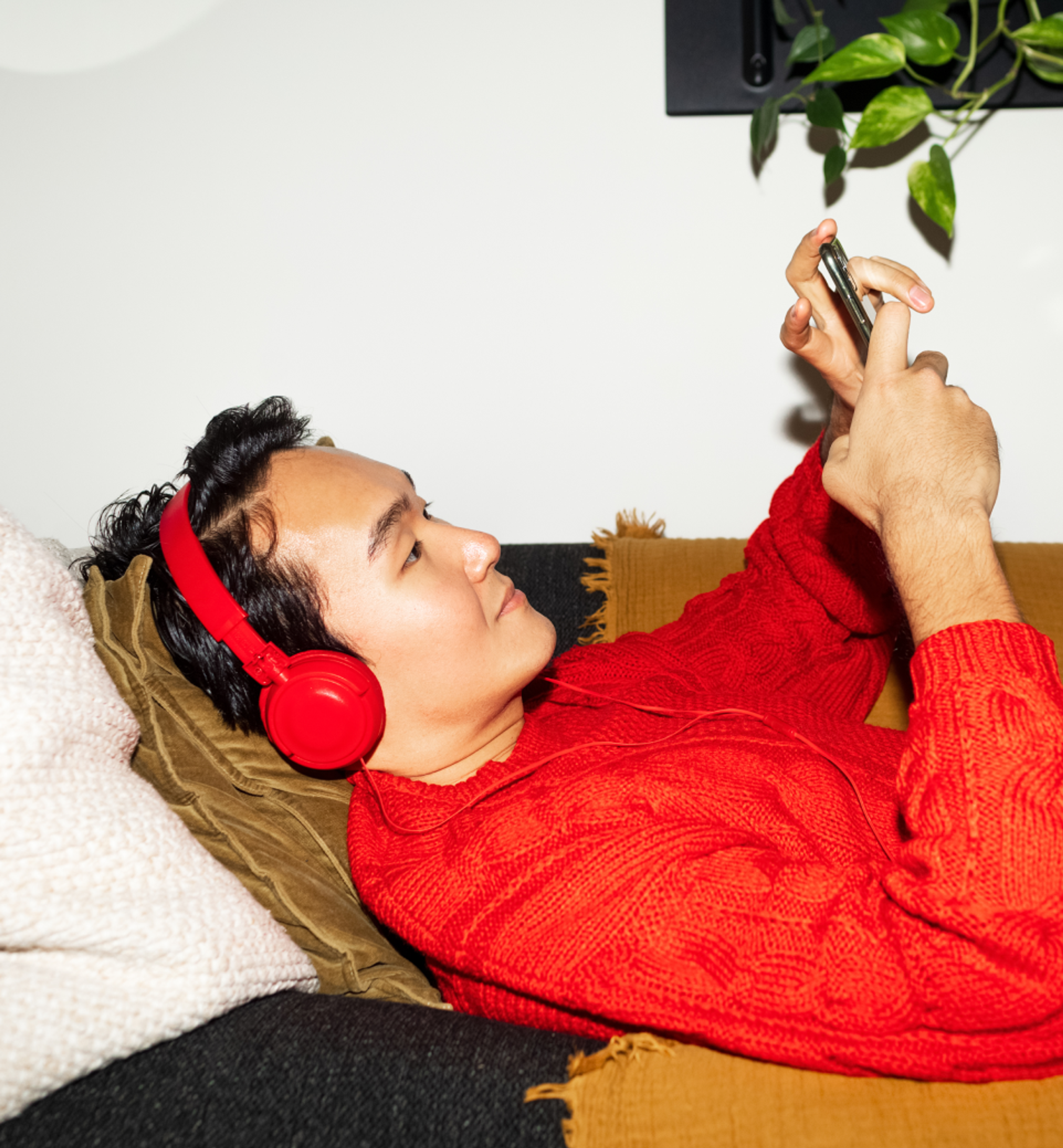 Person lying on a couch wearing bright red headphones and a red sweater, looking at a smartphone held above them.