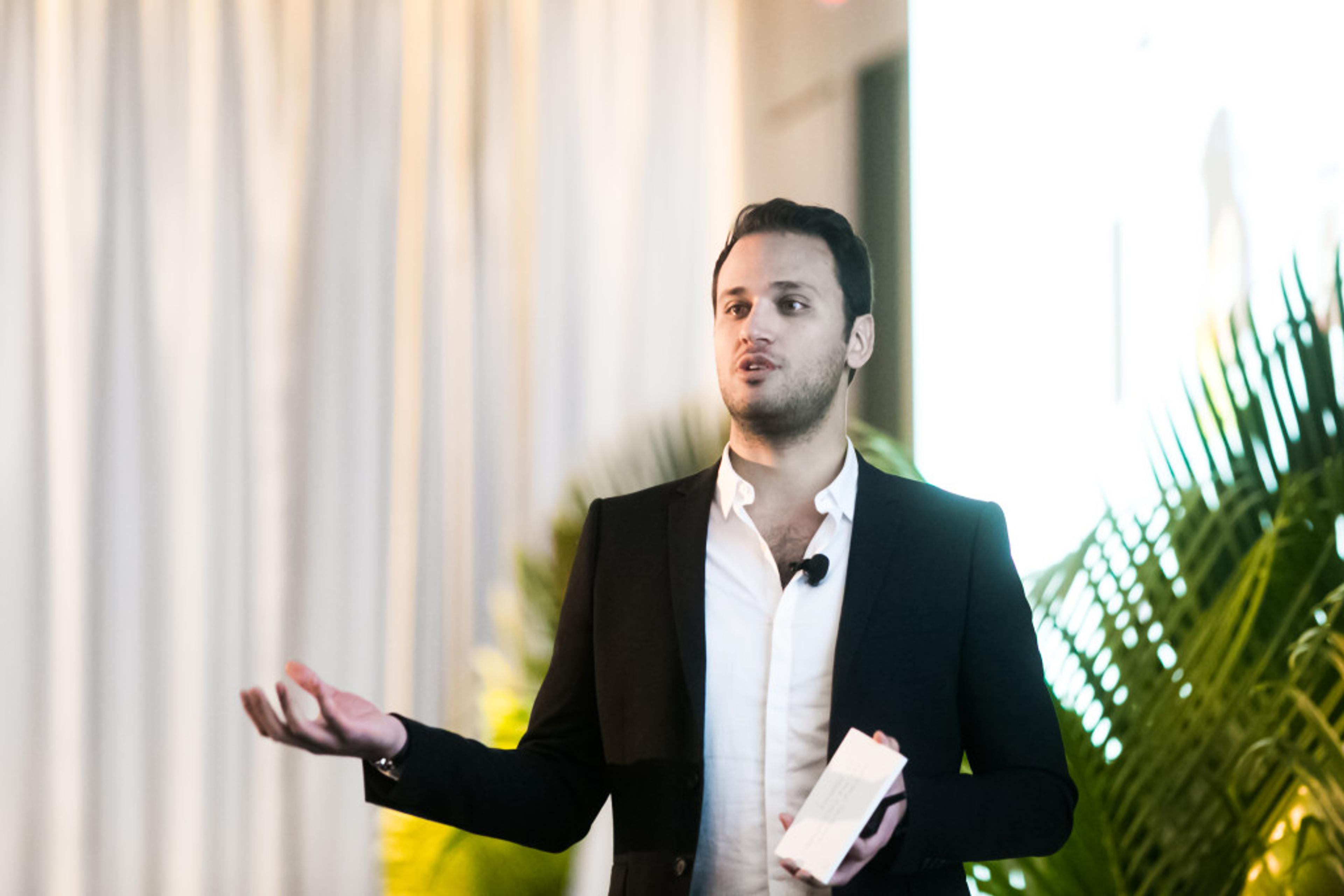 A man in a black suit gestures while speaking at an event, with a microphone attached to his lapel. Green plants are visible in the background.