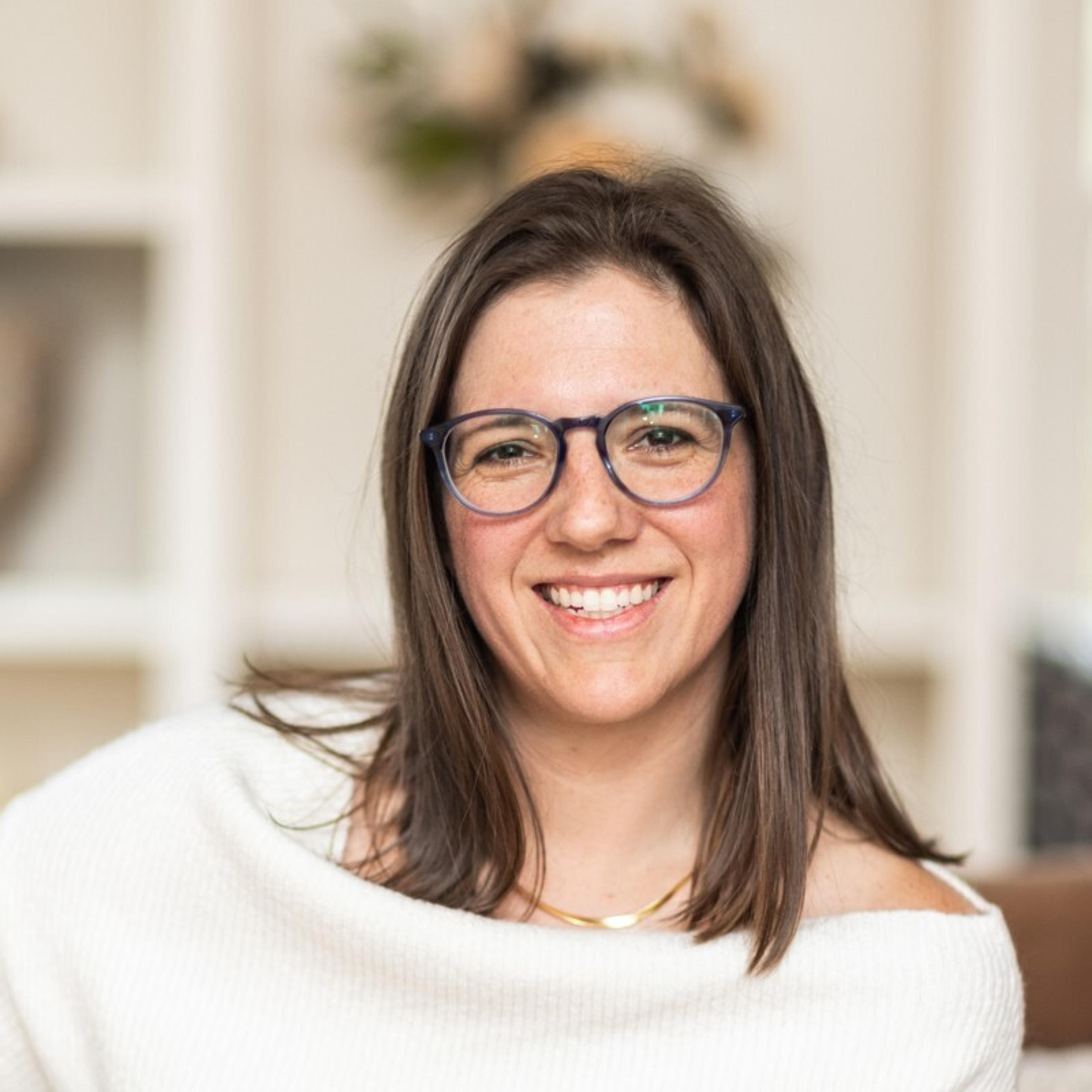A smiling woman with long brown hair and glasses is sitting indoors. She is wearing a white sweater and has a warm, friendly expression.