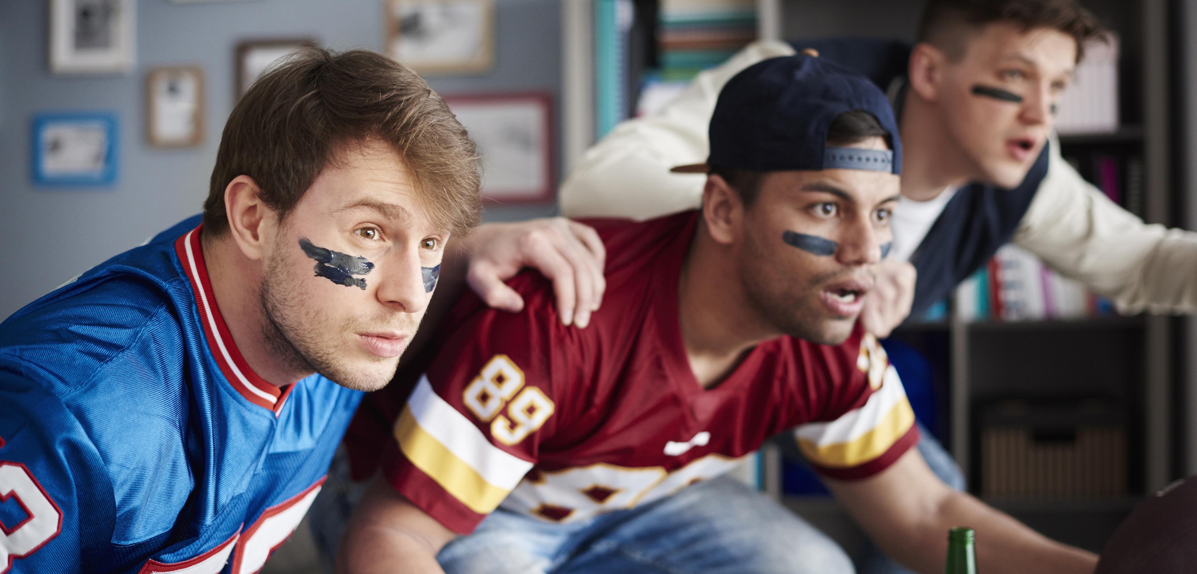 Three young men in football jerseys with eye black intently watch a game.