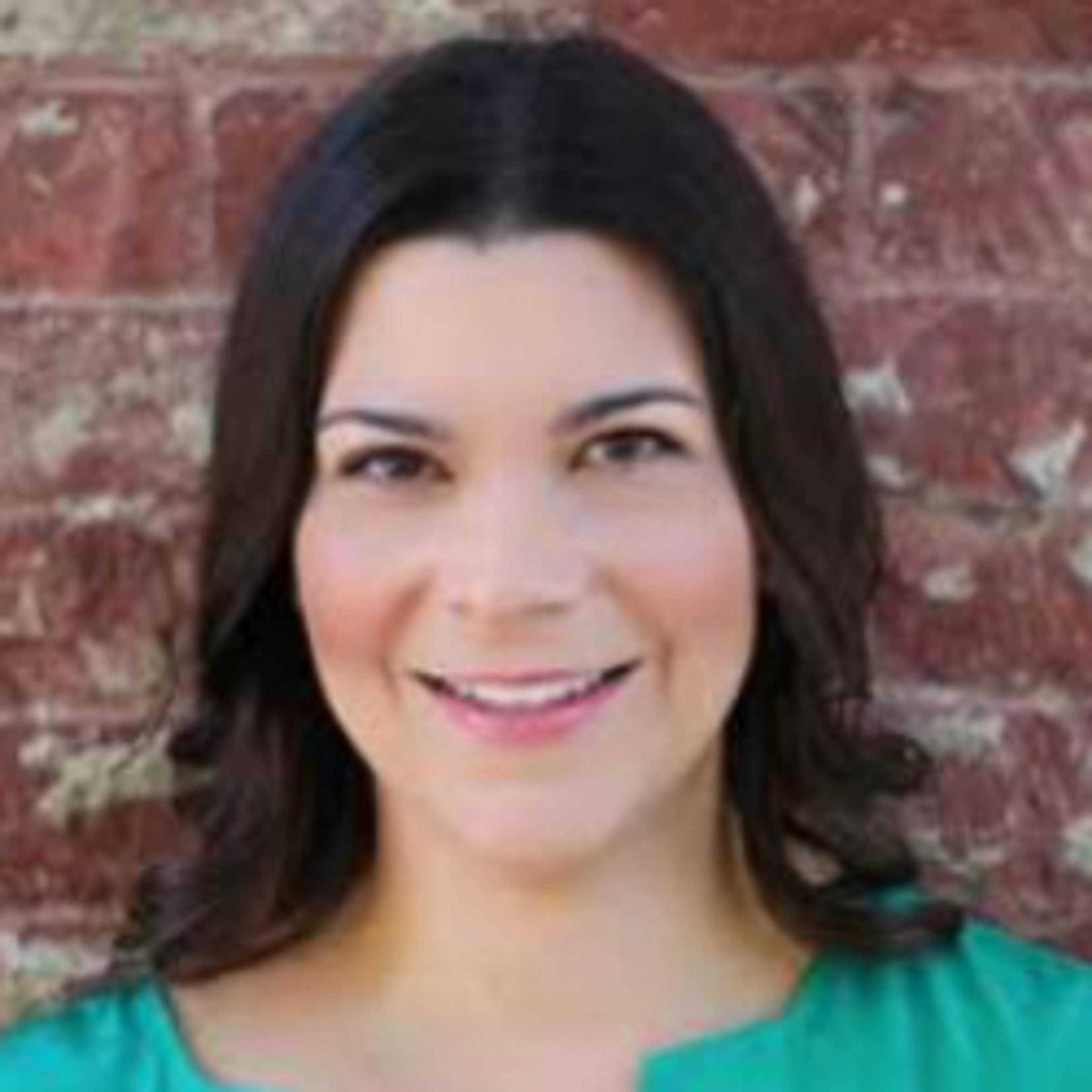 A woman with long dark hair smiles at the camera, wearing a green top against a brick wall background.