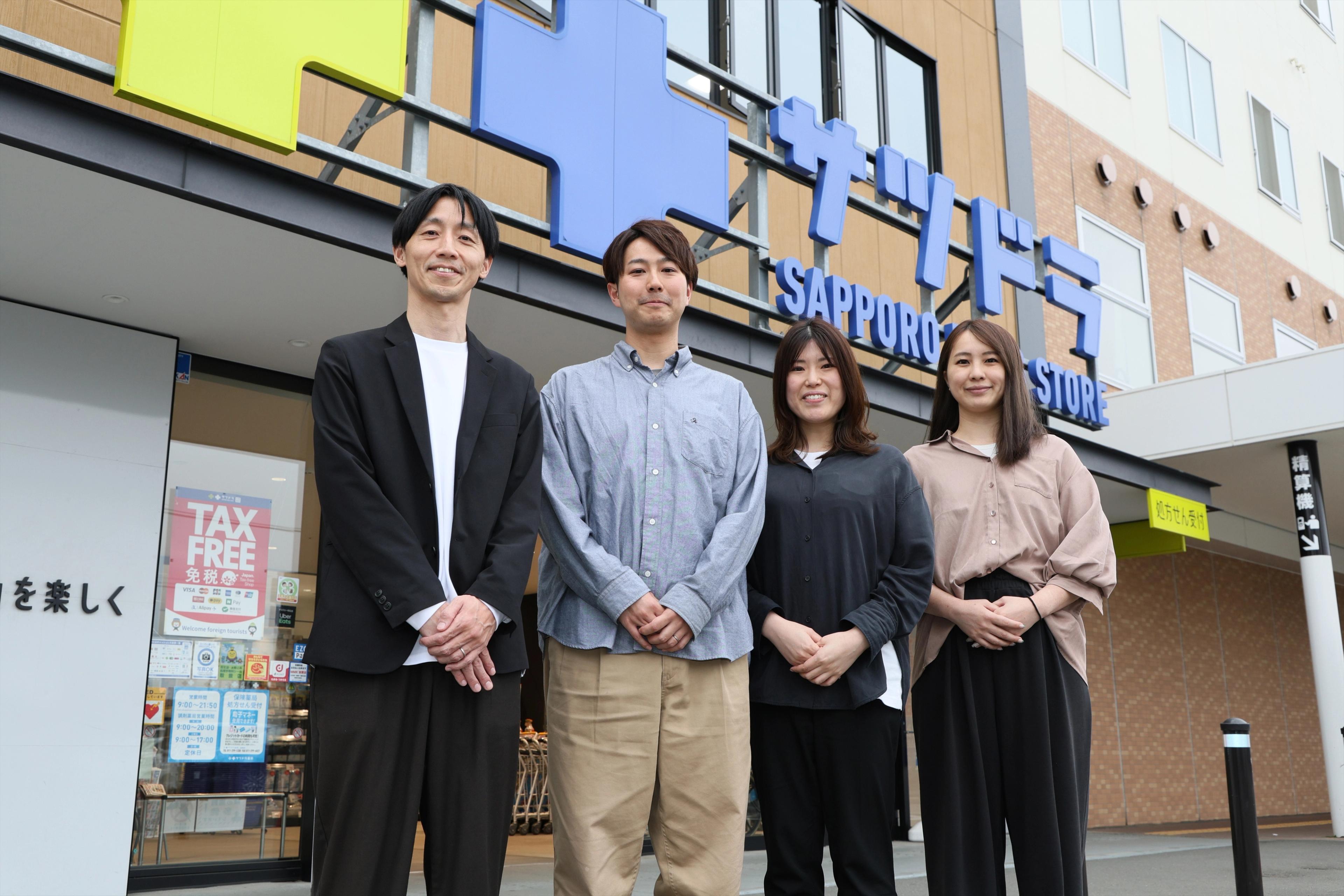 サツドラのインタビューを受けた社員4名がサツドラのロゴのビルの前で笑顔で並んでいる