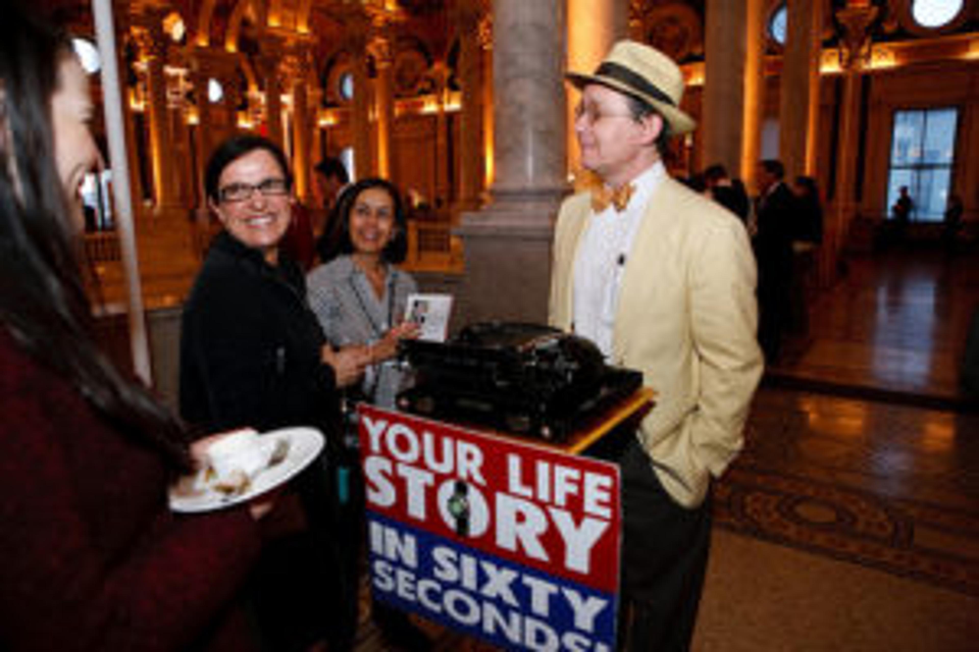 A man in a straw hat stands next to a typewriter, engaging with two women who are smiling. The backdrop features ornate architecture and warm lighting.