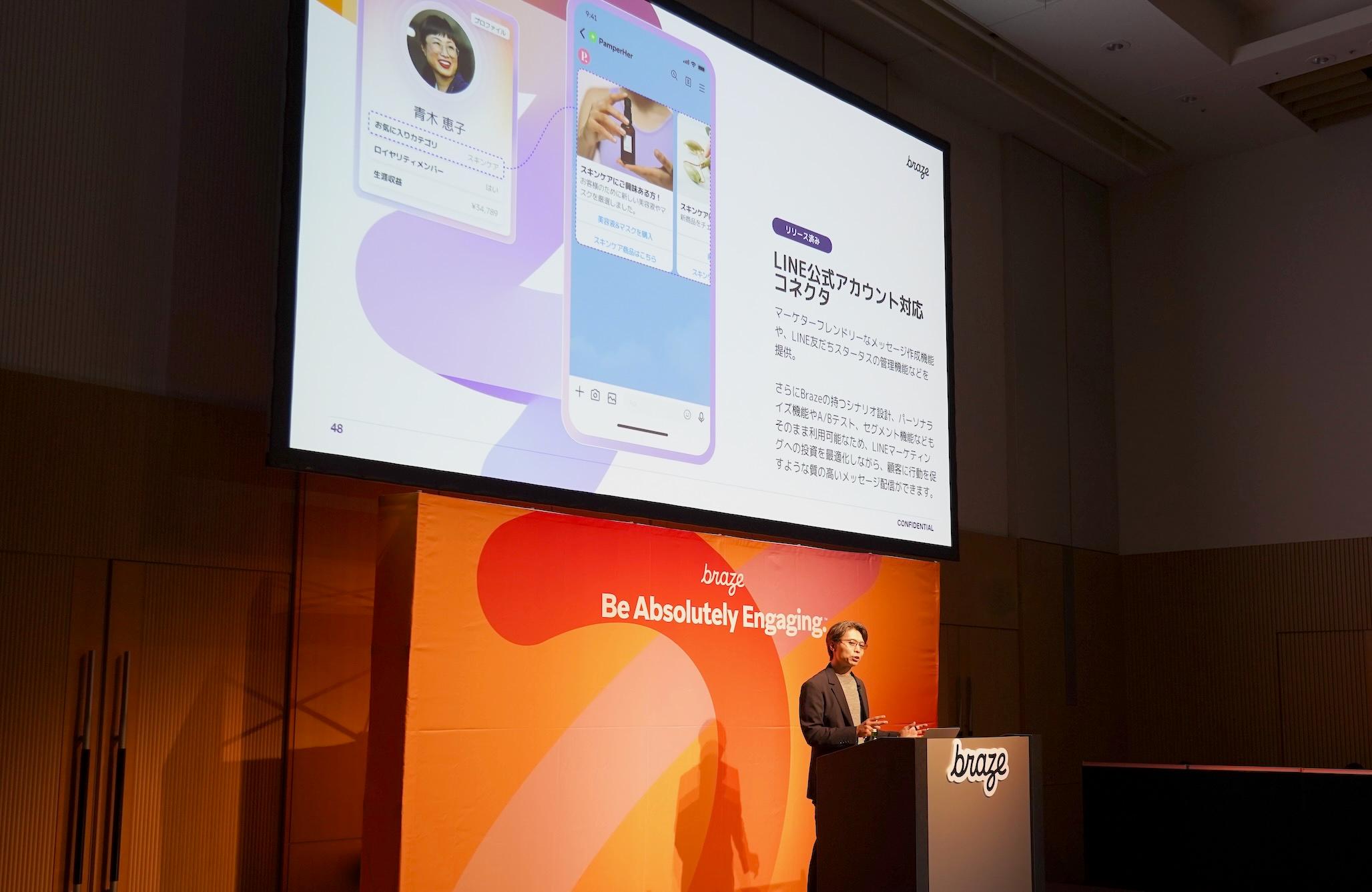 プレゼンテーション中の男性が、LINEアカウントの機能を紹介しています。背景には「Be Absolutely Engaging」のスローガンがあります。