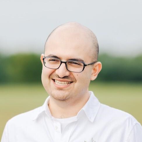 A smiling man with glasses and a bald head stands outdoors, wearing a white shirt. The background features a blurred green landscape.