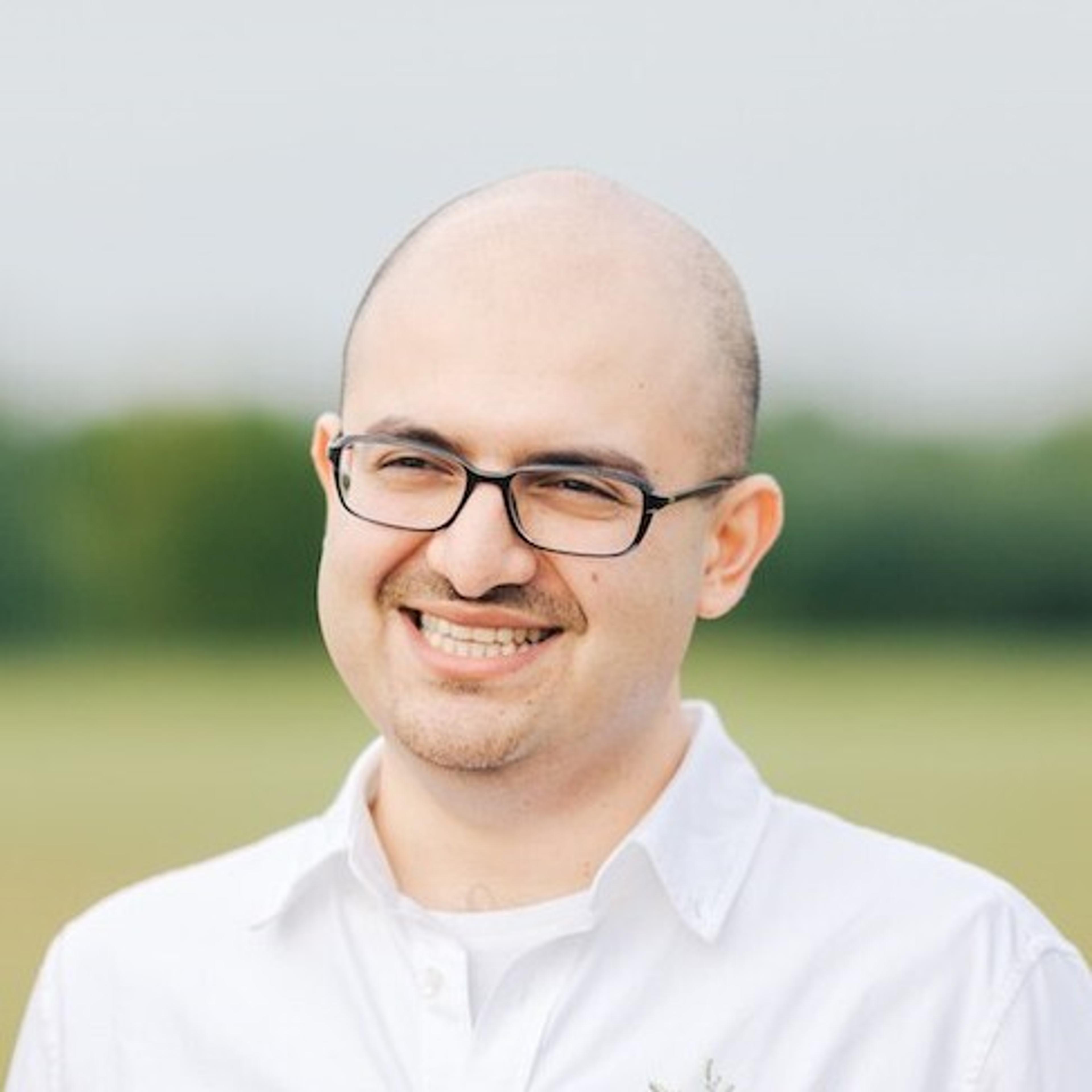 A smiling man with glasses and a bald head stands outdoors, wearing a white shirt. The background features a blurred green landscape.