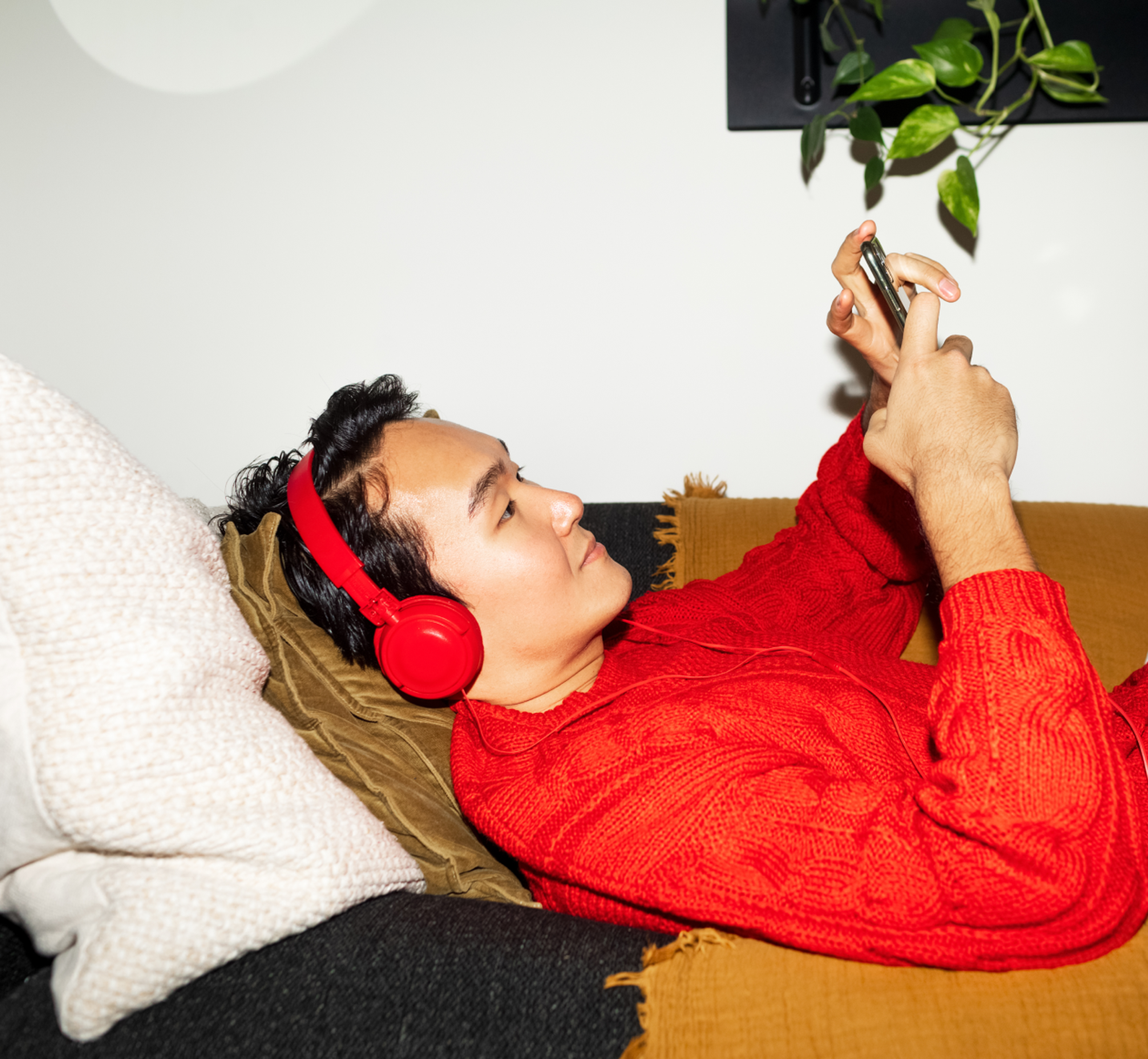 Person lying on a couch in a bright red sweater and matching red headphones, looking at a smartphone held above them.