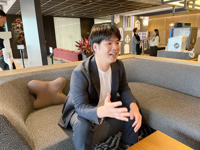 男性がソファに座り、話をしている様子。背景には他の人々やインテリアが見える。