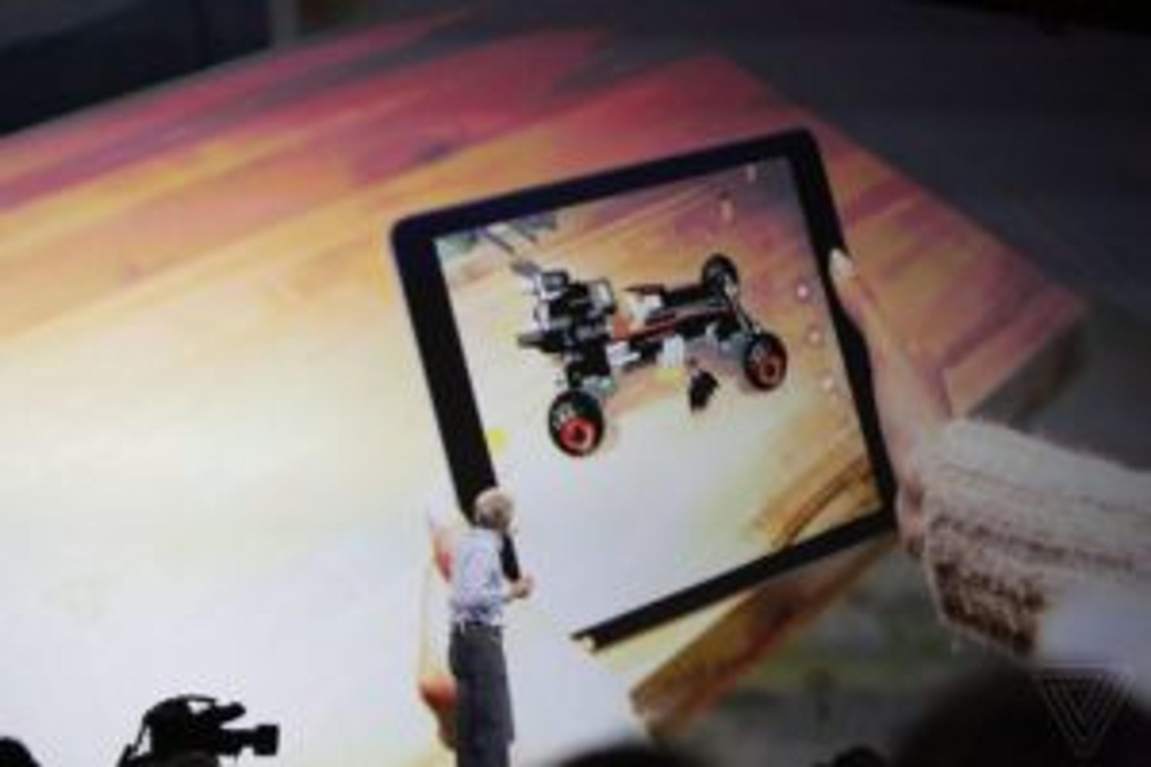 A person stands on stage presenting an augmented reality demonstration on a tablet, showing a 3D model of a toy car. The background features a large screen displaying the same model.