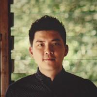A man with short, dark hair stands in front of a blurred green background. He is wearing a dark patterned shirt and looking directly at the camera.