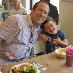 A man and a girl with face paint smile at the camera while sitting at a table with food. The girl is playfully posing with her hand.