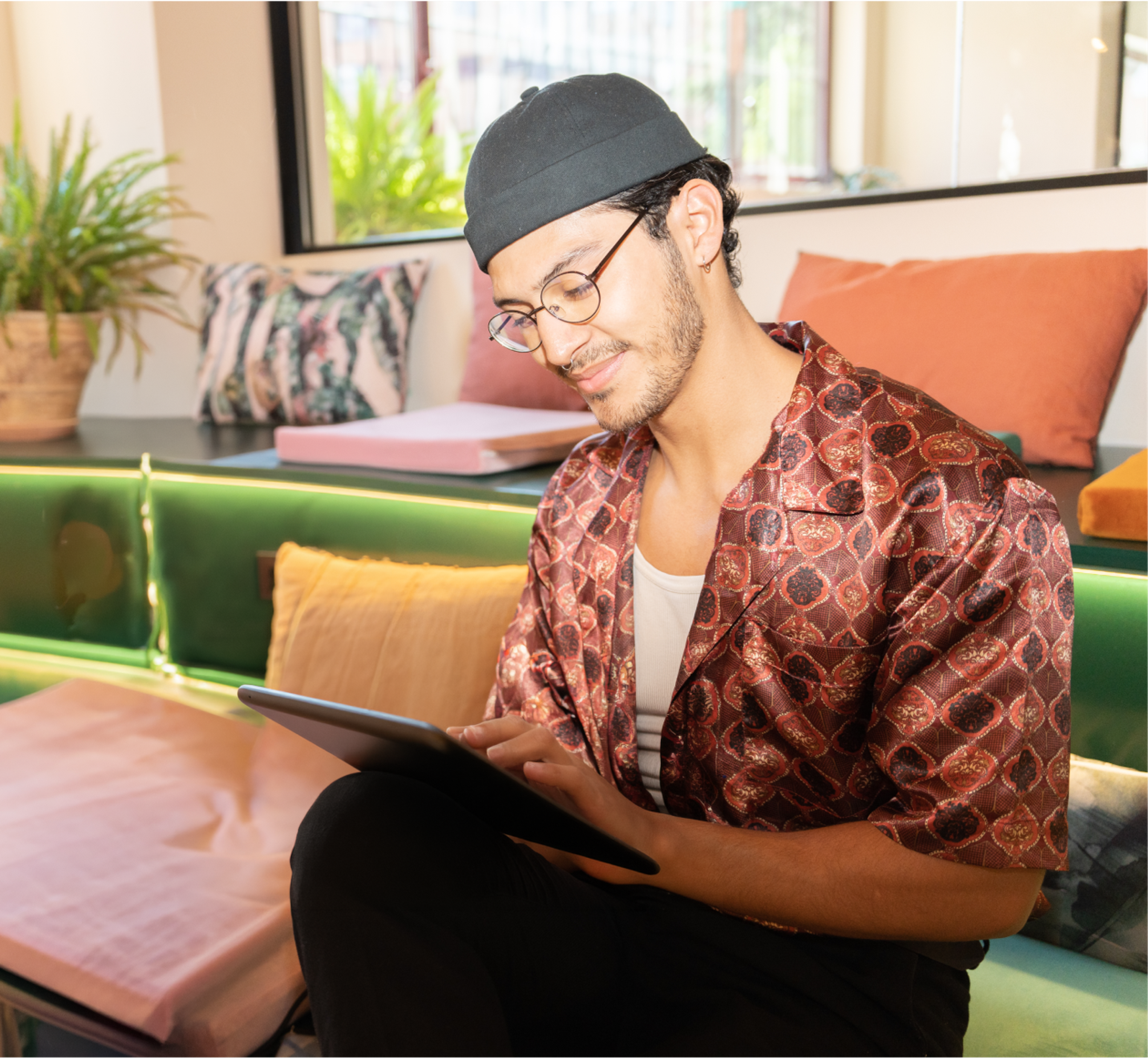 Person wearing glasses and a patterned shirt sits on a colorful couch, smiling while using a tablet in a bright, modern lounge.