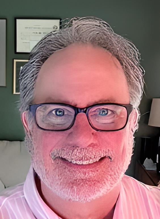 A man with gray hair and glasses smiles at the camera, sitting in a well-lit room with framed documents in the background. He has a light pink shirt on.