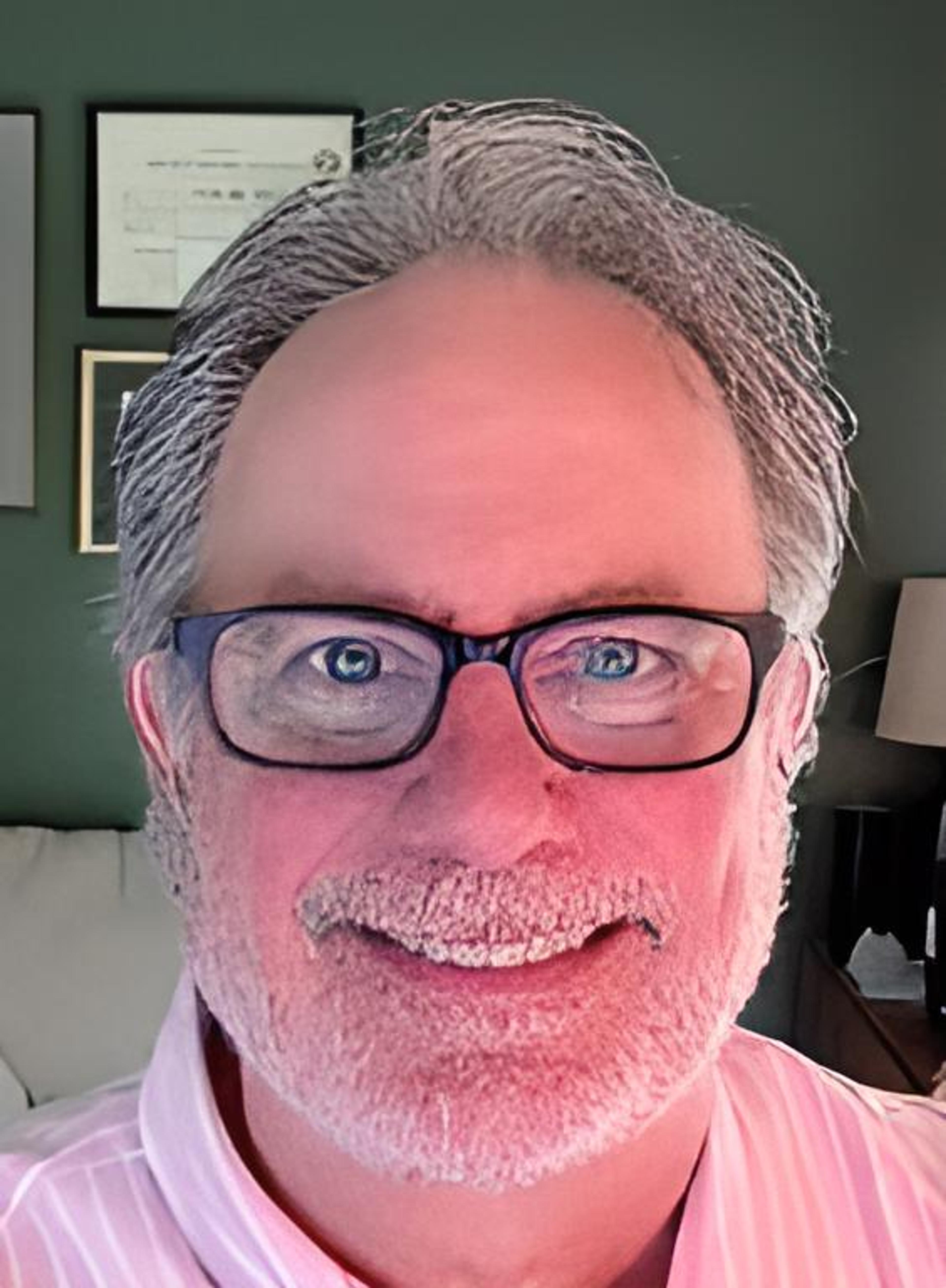 A man with gray hair and glasses smiles at the camera, sitting in a well-lit room with framed documents in the background. He has a light pink shirt on.