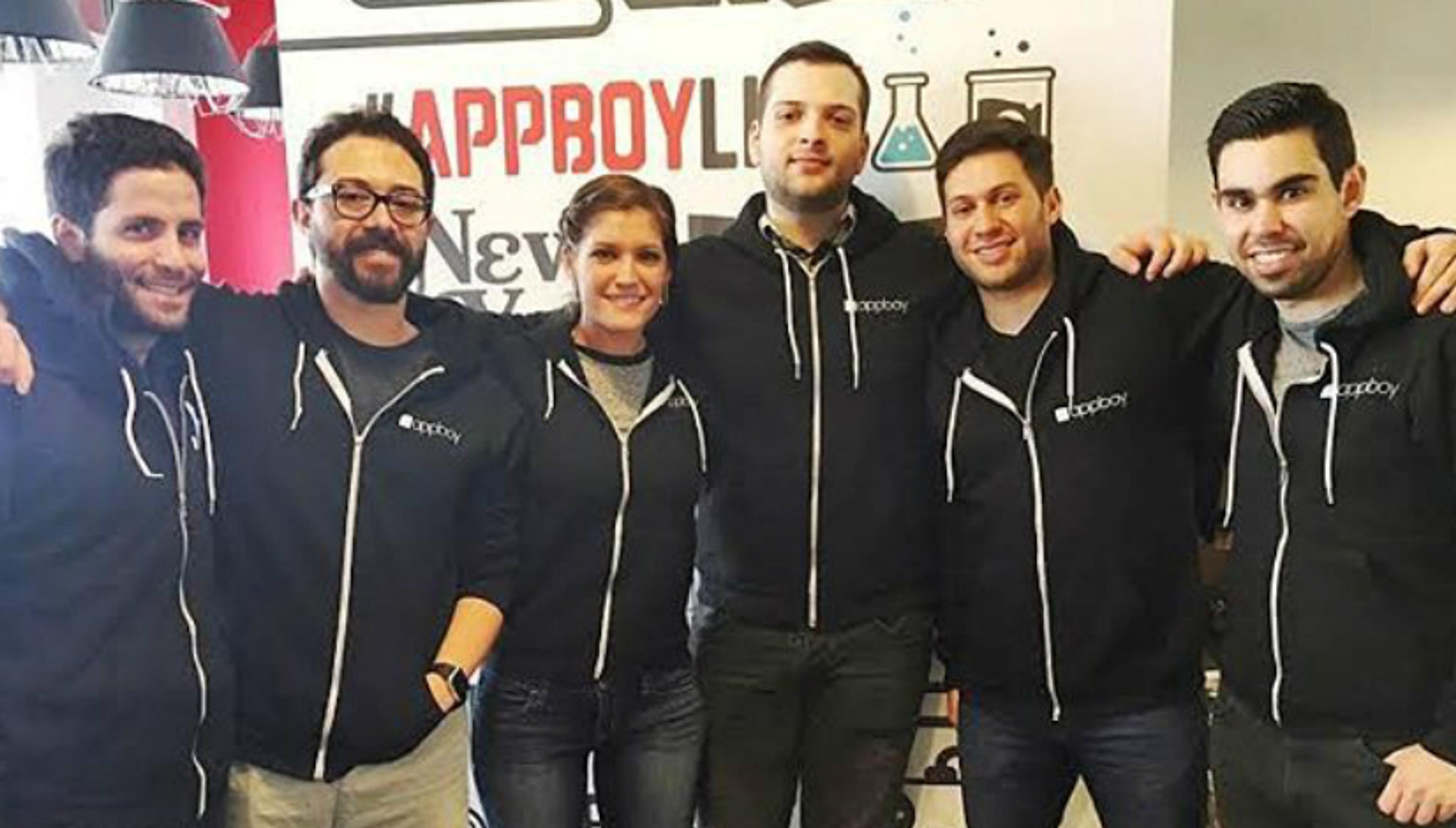A group of six people wearing matching black hoodies stands together, smiling in front of a colorful backdrop with the text "#AppBoyLab.