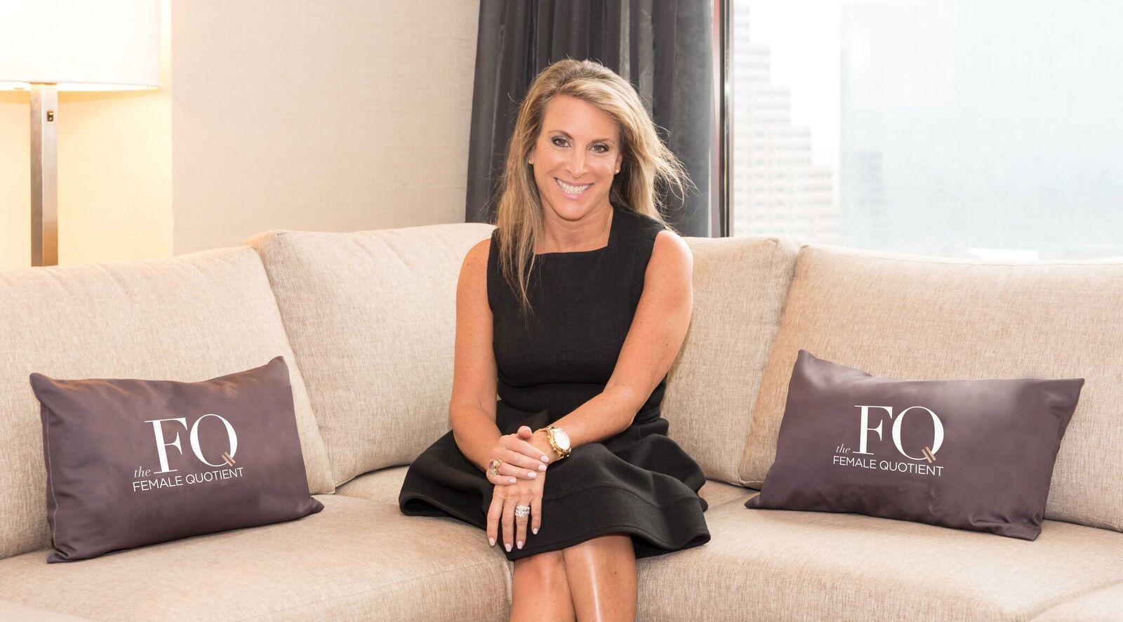 A woman in a black dress sits on a beige couch, smiling at the camera. Two decorative pillows with the logo "FQ the Female Quotient" are placed on the couch.