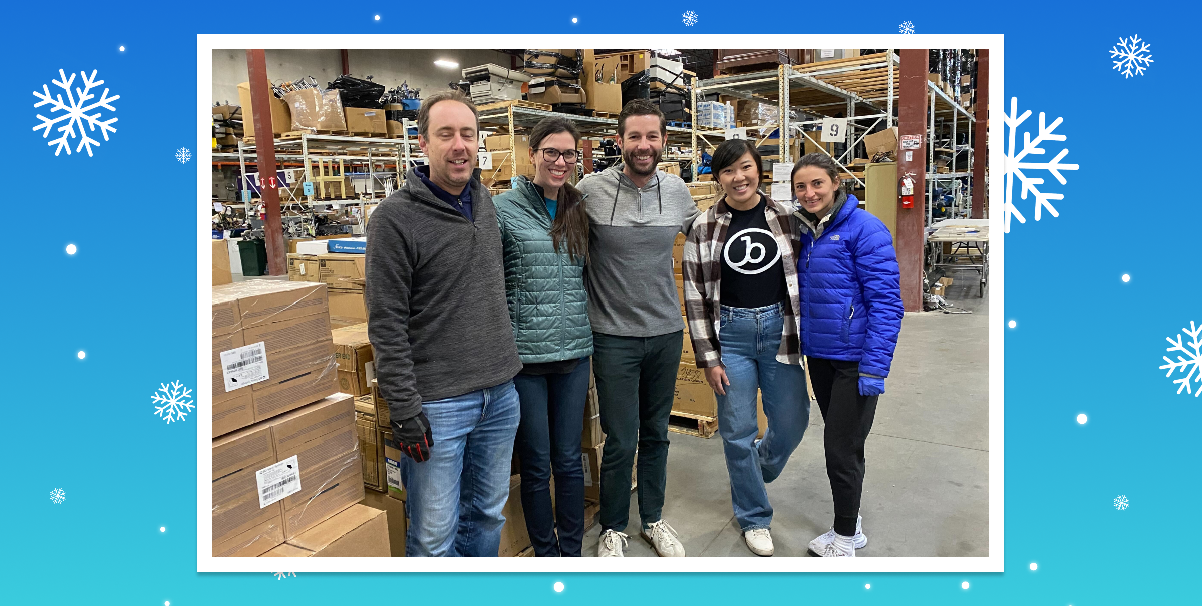 A group of five people stands in a warehouse surrounded by boxes, smiling at the camera. They are dressed in casual winter clothing.
