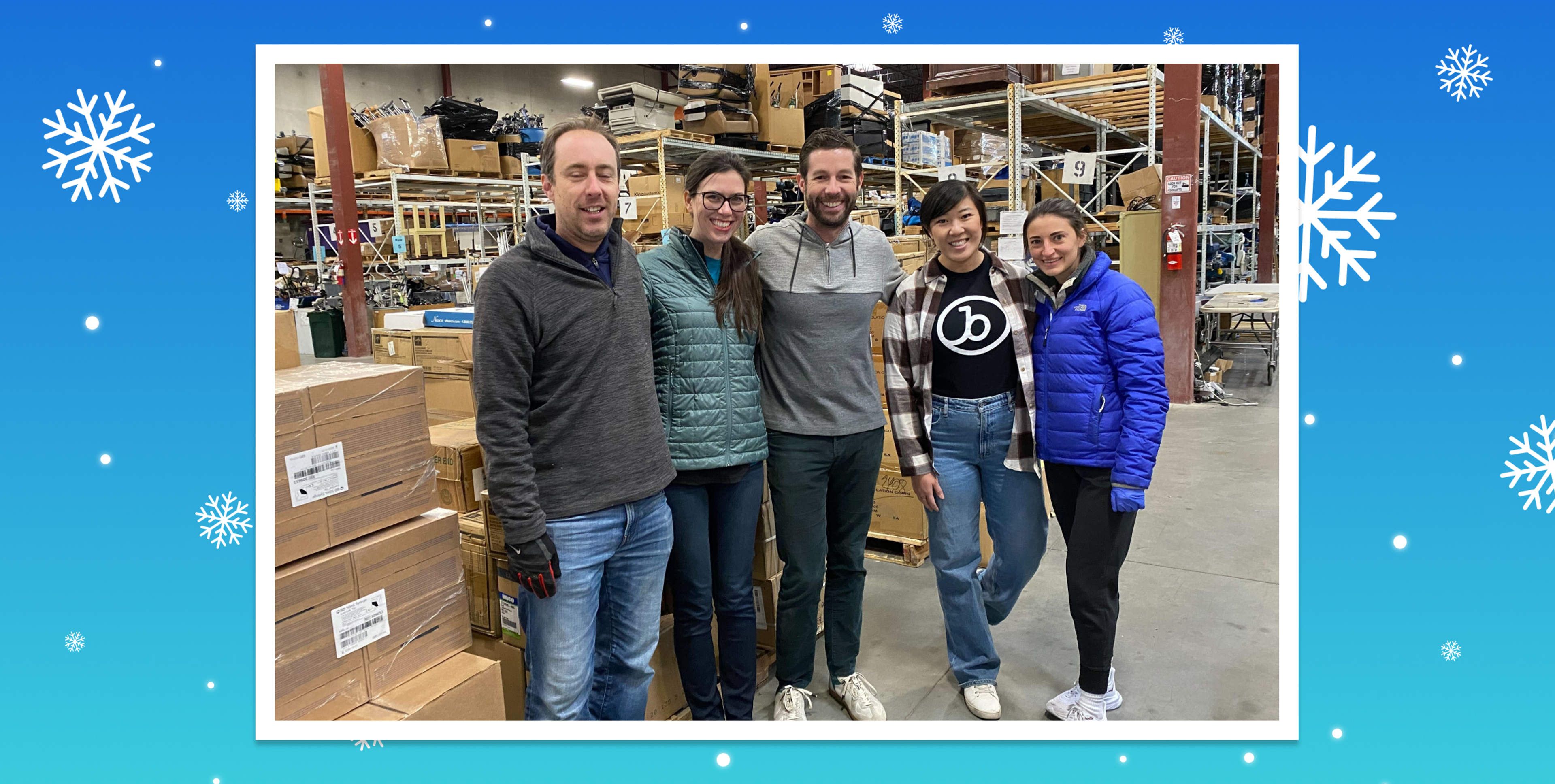 A group of five people stands in a warehouse surrounded by boxes, smiling at the camera. They are dressed in casual winter clothing.