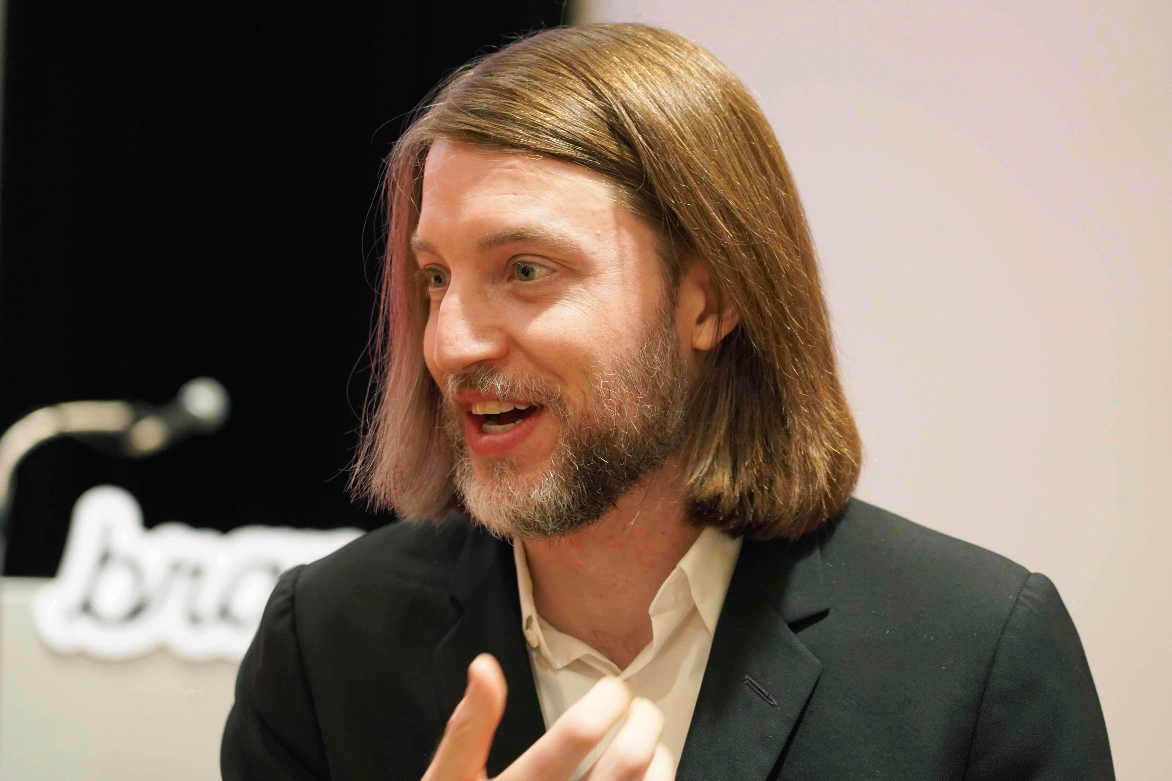 a man with long hair and a beard is wearing a suit and talking into a microphone .