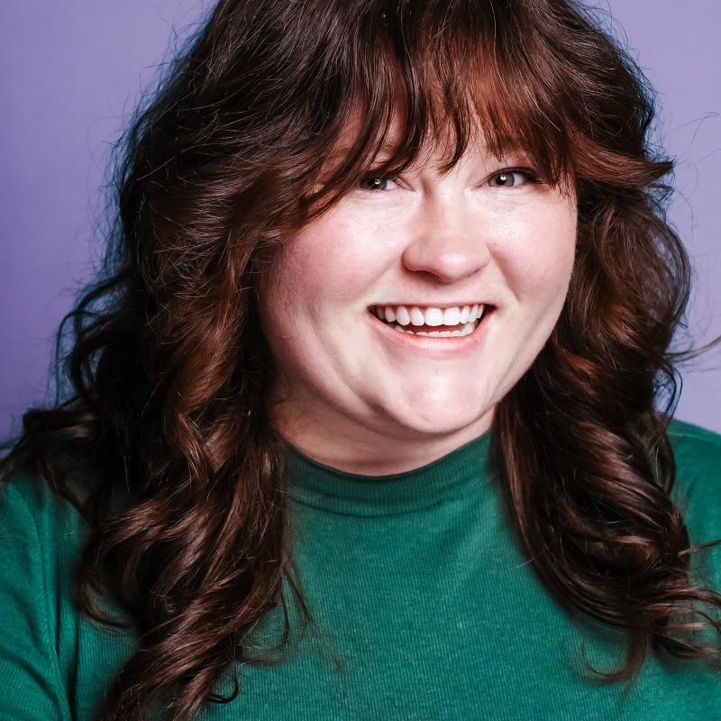 A smiling person with long, wavy brown hair wearing a green shirt against a purple background. The expression is cheerful and friendly.
