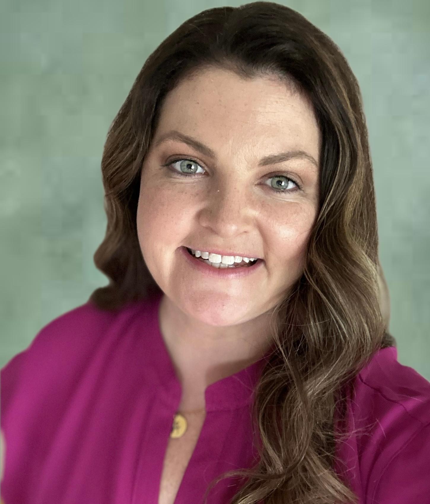 A woman with long, wavy brown hair smiles at the camera, wearing a bright pink blouse. She has blue eyes and a warm expression.
