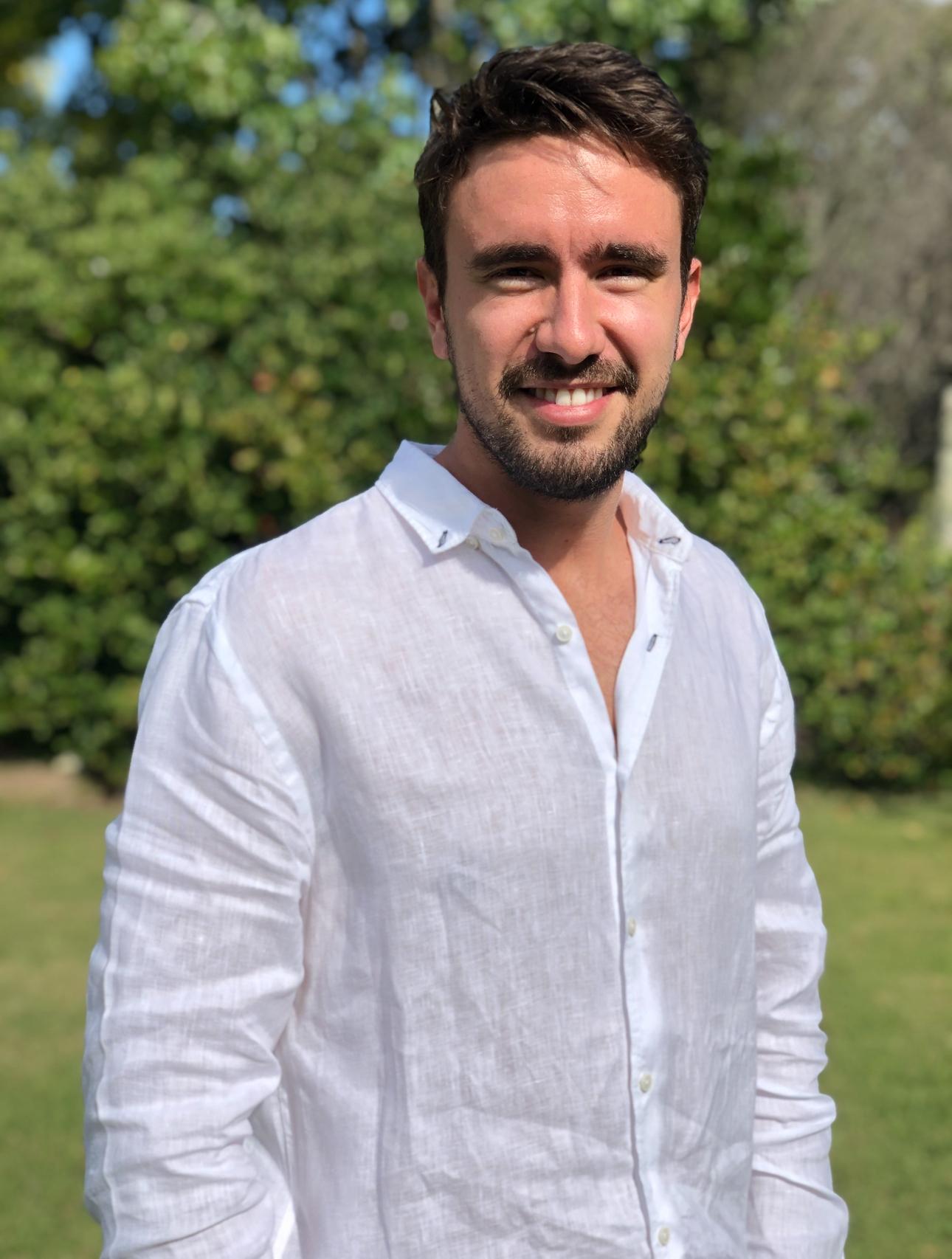 Hombre sonriente con camisa blanca de lino, de pie en un jardín con árboles al fondo.