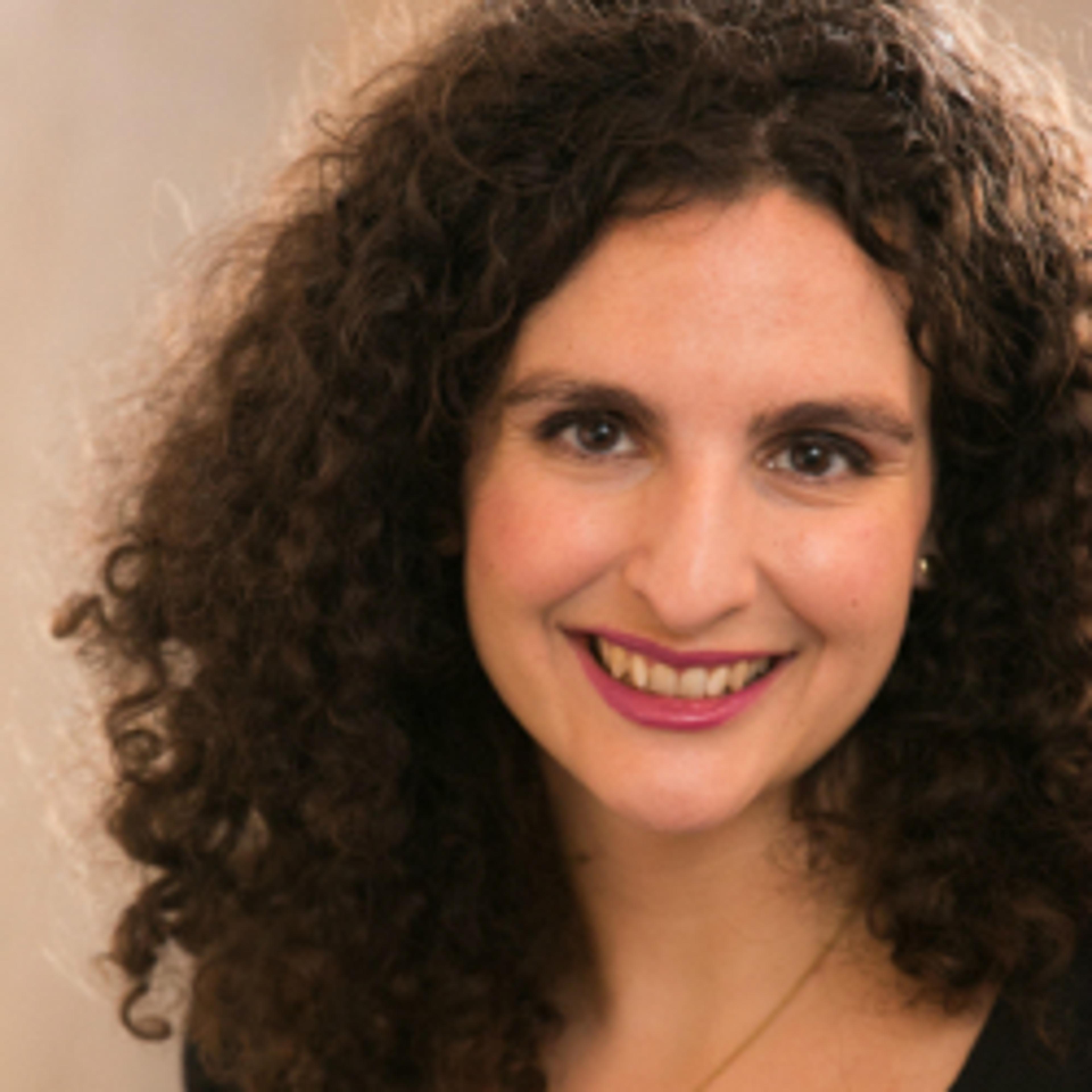 A smiling woman with curly hair and a warm expression. She is wearing a light-colored top and has a subtle necklace.