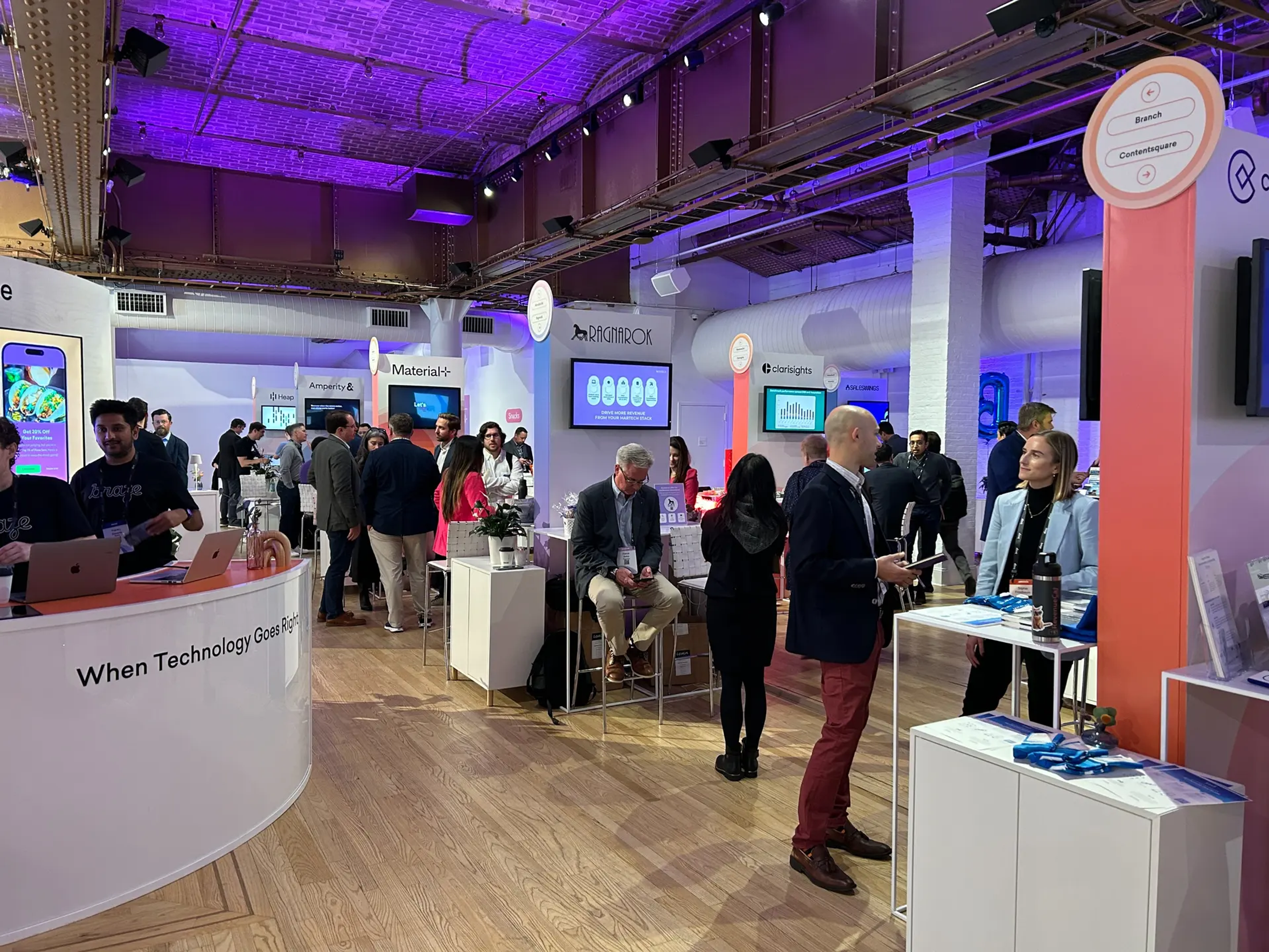 Una feria tecnológica con varias estaciones de exhibición. Personas interactuando y conversando en un ambiente moderno y iluminado.