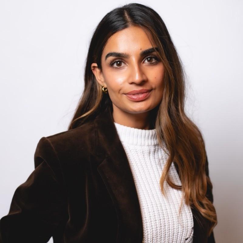 Professional headshot of a woman with long wavy hair wearing a white knit sweater and dark blazer, smiling slightly against a plain light background.