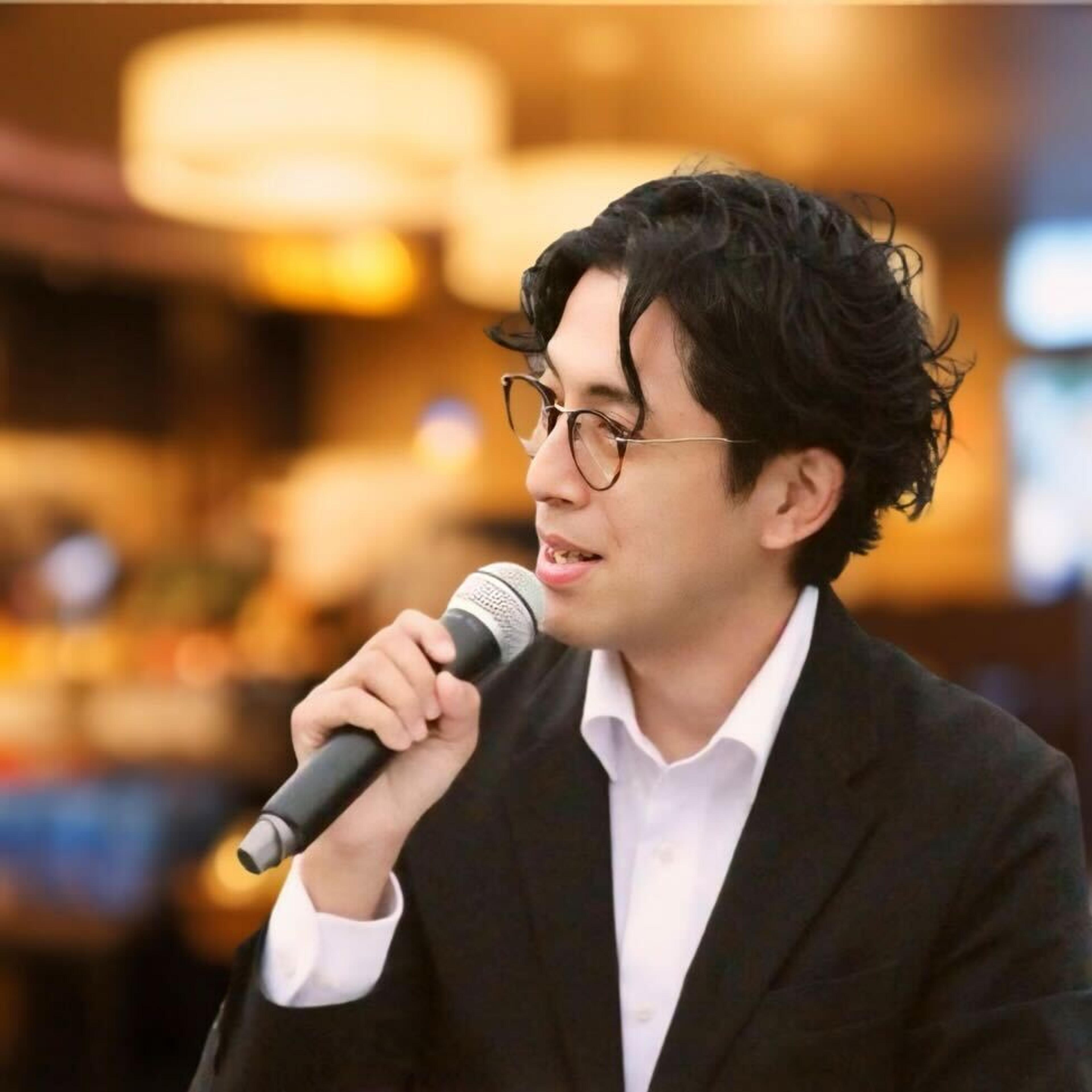 マイクを持って話す男性が、カジュアルな雰囲気の場所でインタビューを受けています。背景にはぼんやりとした照明があります。