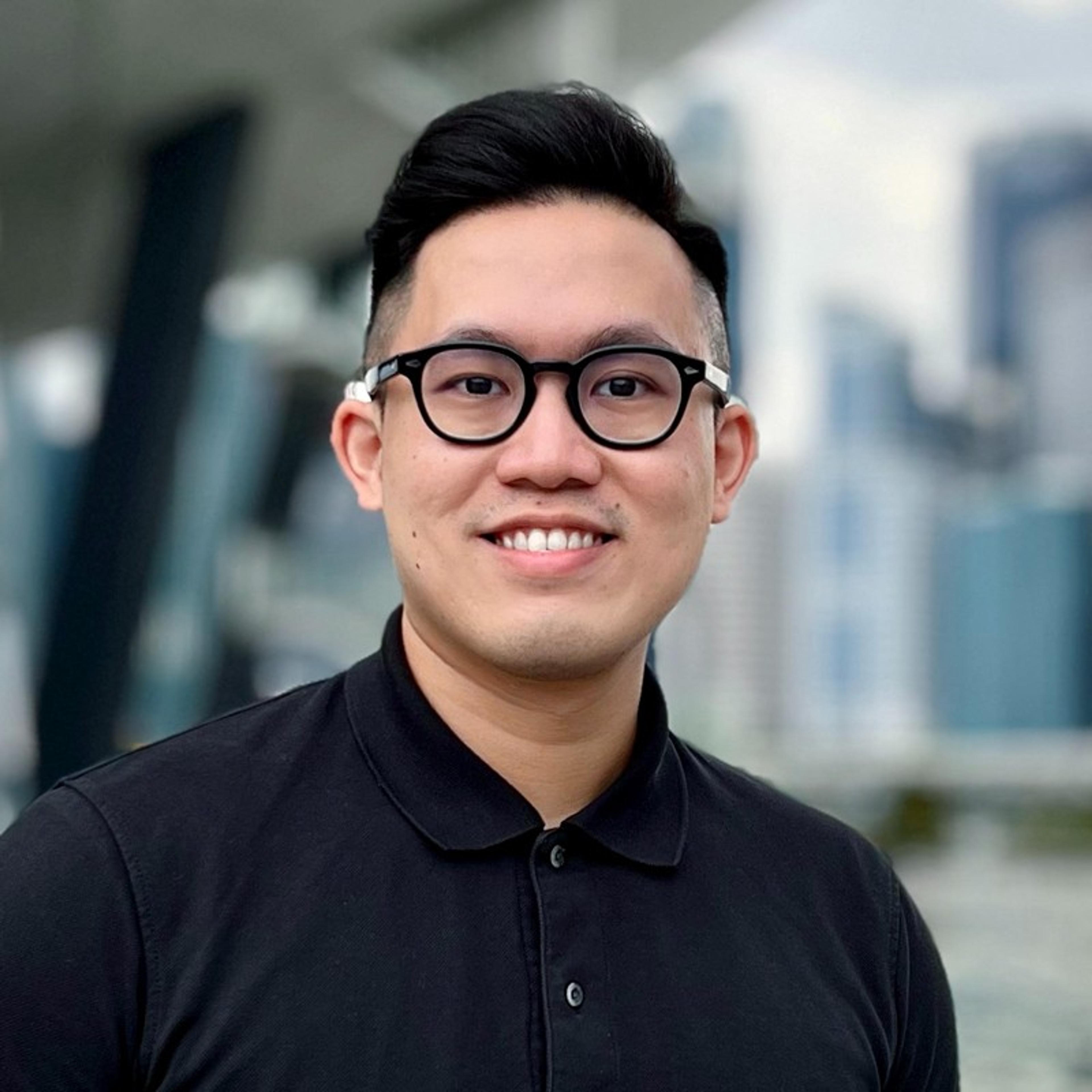 A young man with glasses and short hair smiles at the camera, standing outdoors with a city skyline in the background. He is wearing a black polo shirt.
