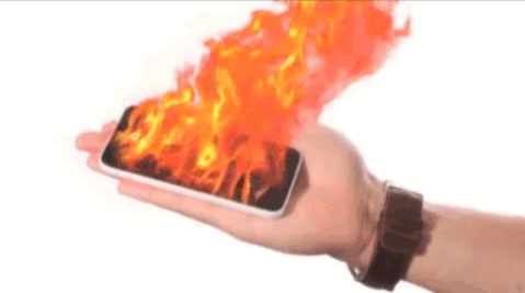A hand holds a smartphone that appears to be engulfed in flames. The flames are bright orange and red, creating a dramatic effect.