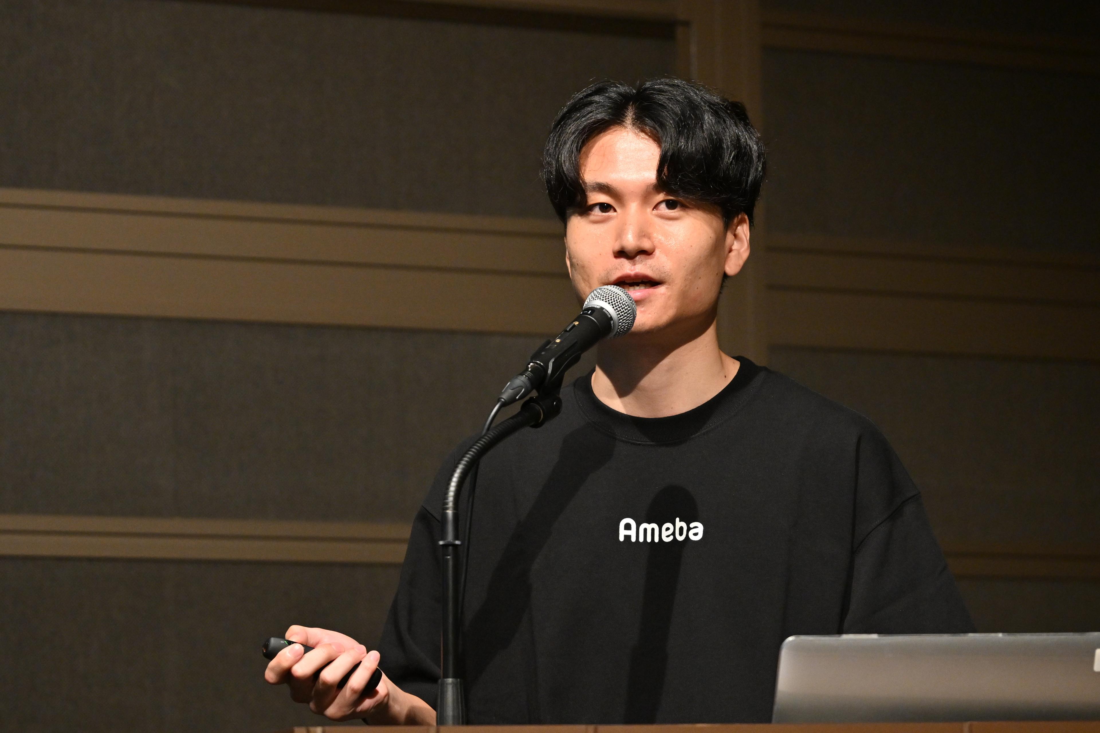 男性がマイクを持ってプレゼンテーションを行っています。Tシャツには「Ameba」と書かれています。